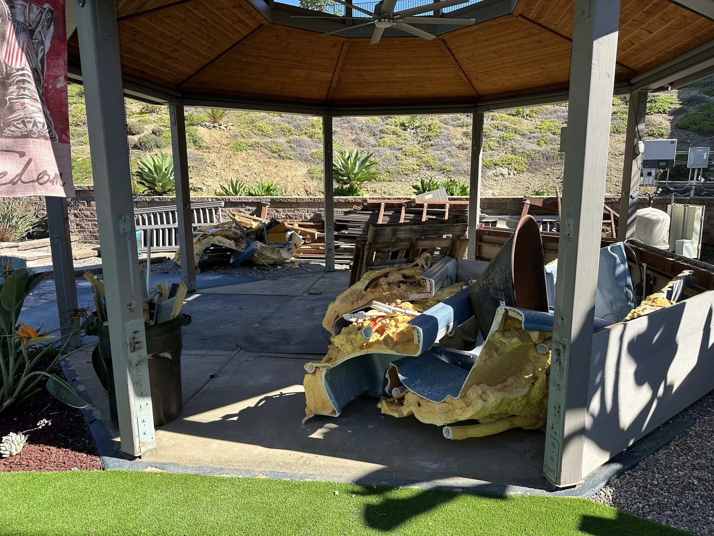 A partially assembled outdoor gazebo with a wooden interior ceiling surrounded by a small garden with plants and rocks. The gazebo contains scattered debris and construction materials, including insulation foam, pipes, and miscellaneous items.