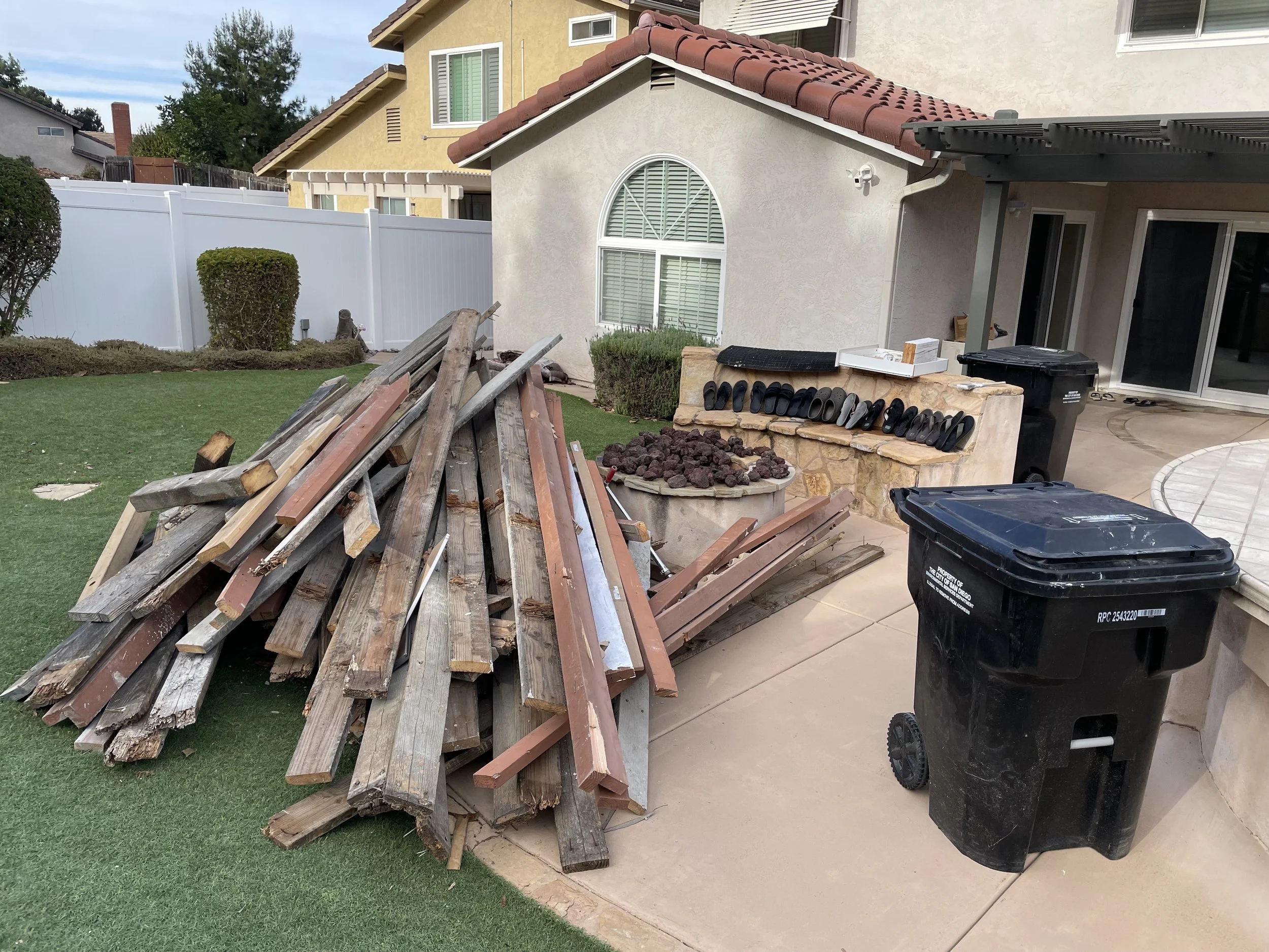 Pile of wooden planks and scrap wood in a backyard next to a pile of rocks, with patio area, trash bins, and houses in the background.