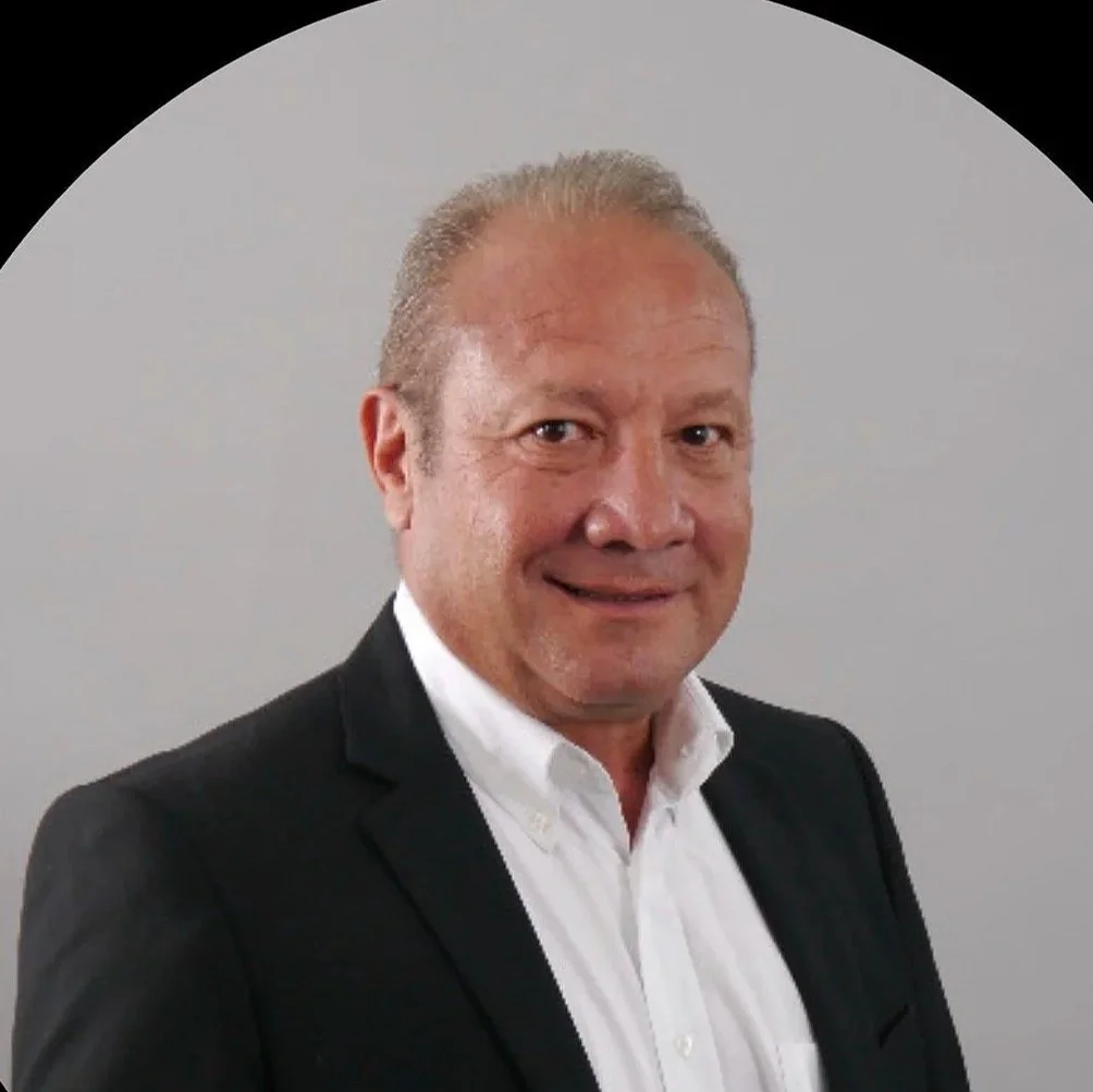 Un homme d'affaires porte un costume noir et une chemise blanche, souriant devant un fond gris clair.