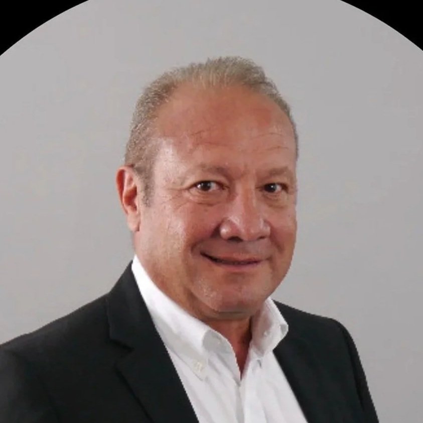 Un homme d'affaires porte un costume noir et une chemise blanche, souriant devant un fond gris clair.