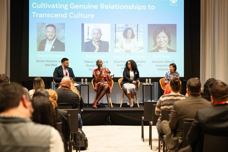 Four panelists seated on stage participating in a discussion at a conference, with a large screen behind them displaying event title and speaker photos. Audience members are watching.