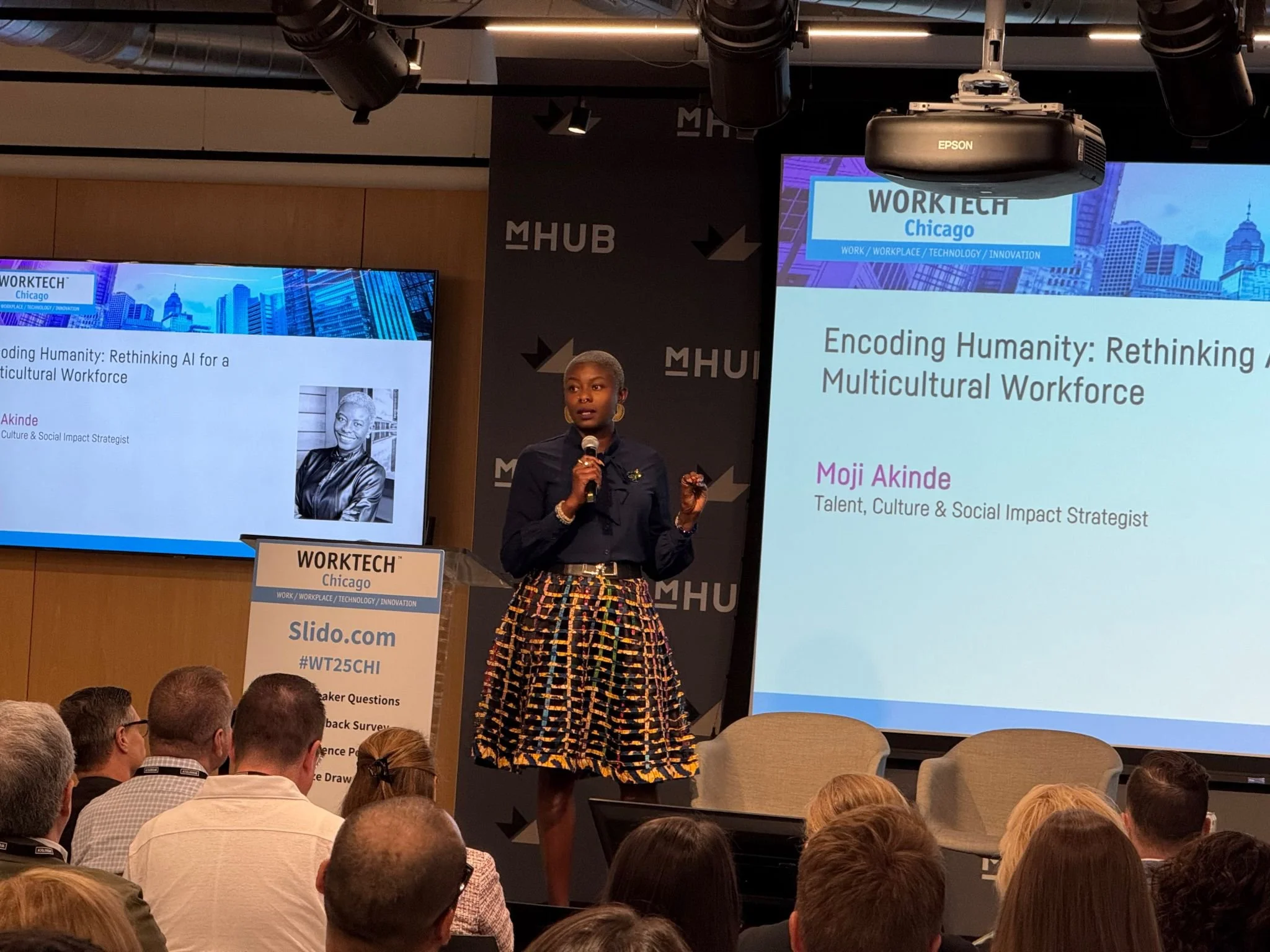 A woman presenting at a conference called WORKTECH Chicago, standing on a stage holding a microphone. Two large screens behind her display her presentation titled 'Encoding Humanity: Rethinking AI for a Multicultural Workforce' with her name, Moji Akinde, and her title as Talent, Culture & Social Impact Strategist. The stage has a podium with a sign for WORKTECH Chicago and some people in the audience.