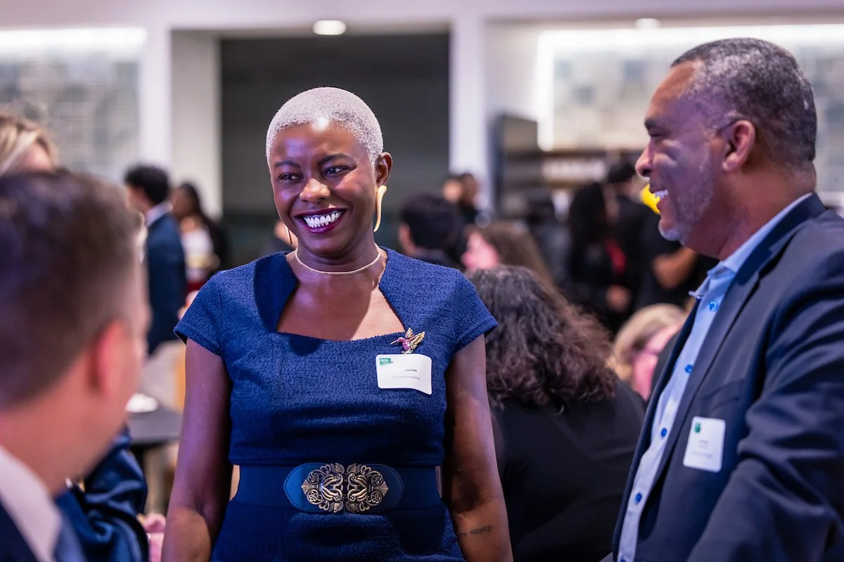 Two people smiling and talking at a professional networking event, with a woman wearing a navy blue dress and a man in a suit, in a crowded indoor space.