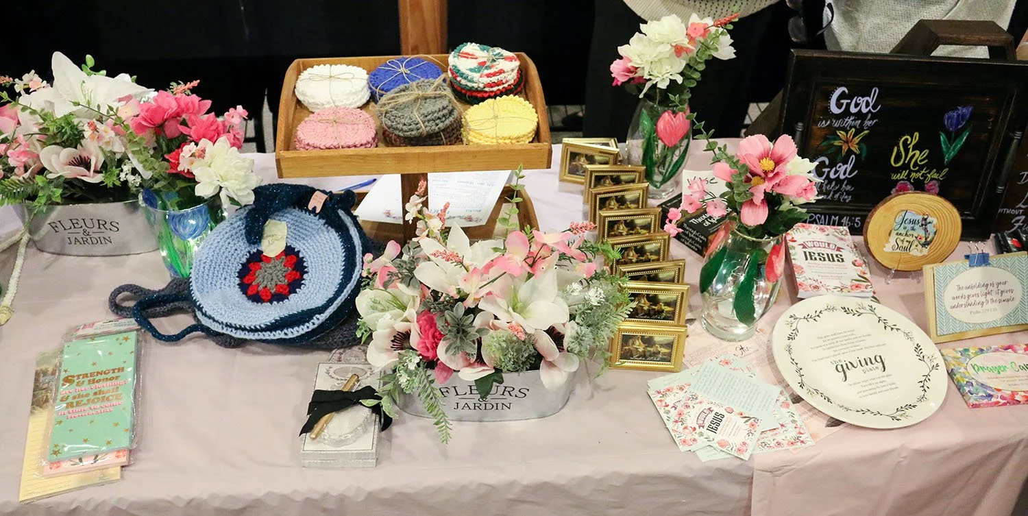 A table decorated with pink flowers in vases, framed photos, religious signs, crocheted items, a round knitted bag, gift cards, and decorative plates with inspirational phrases, all themed around faith and encouragement.