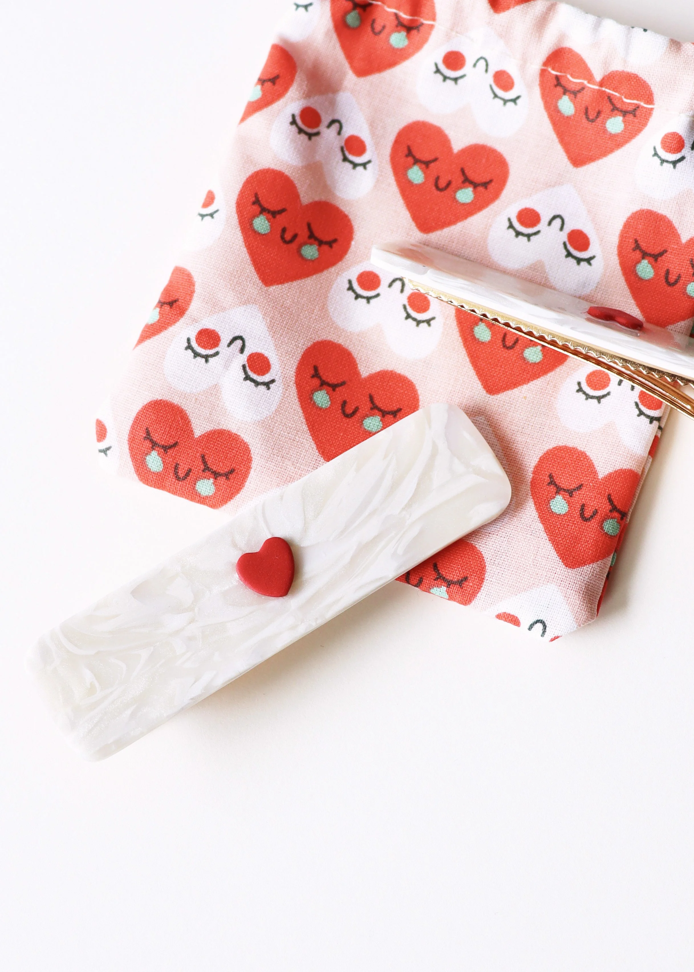 Une pince à cheveux blanche avec un cœur rouge en relief, posé sur un pochon en tissu à motif de cœurs rouges avec des visages souriants et des yeux fermés, le tout sur un fond blanc.