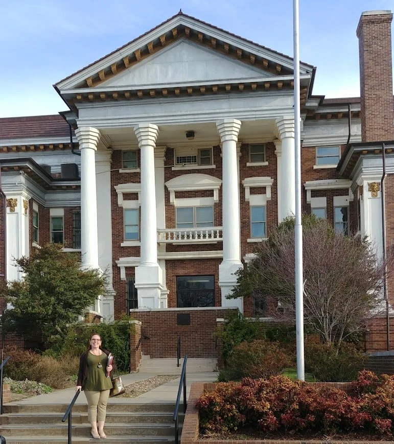 An attorney standing on steps in front of a large, historic brick Texas courthouse with white columns and trim, trees, shrubs, and a flagpole.