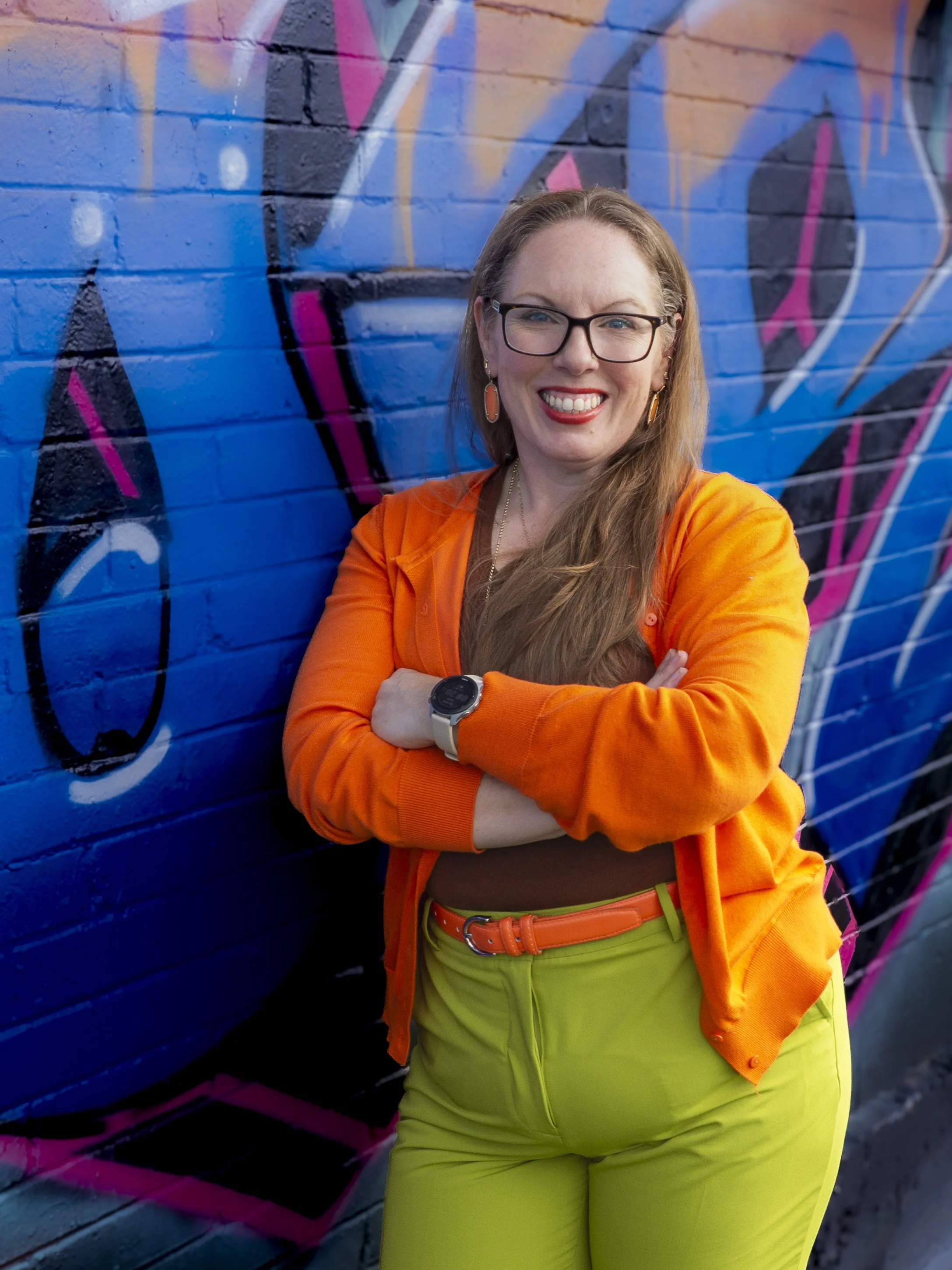 A smiling attorney with glasses, brown hair, and earrings stands next to a colorful graffiti wall. She is wearing an orange jacket, lime green pants, a brown top, and an orange belt, with her arms crossed.