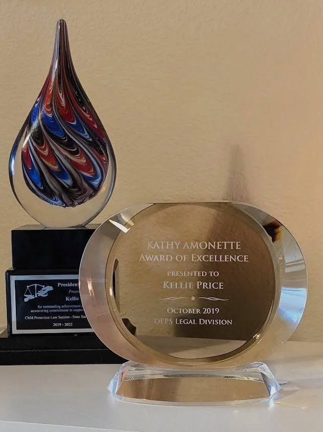 A glass award with the text 'Kathy Amonette Award of Excellence presented to Kellie Price October 2019 DFS Legal Division' and a colorful glass award with swirling red, blue, black, and silver colors on a black base.