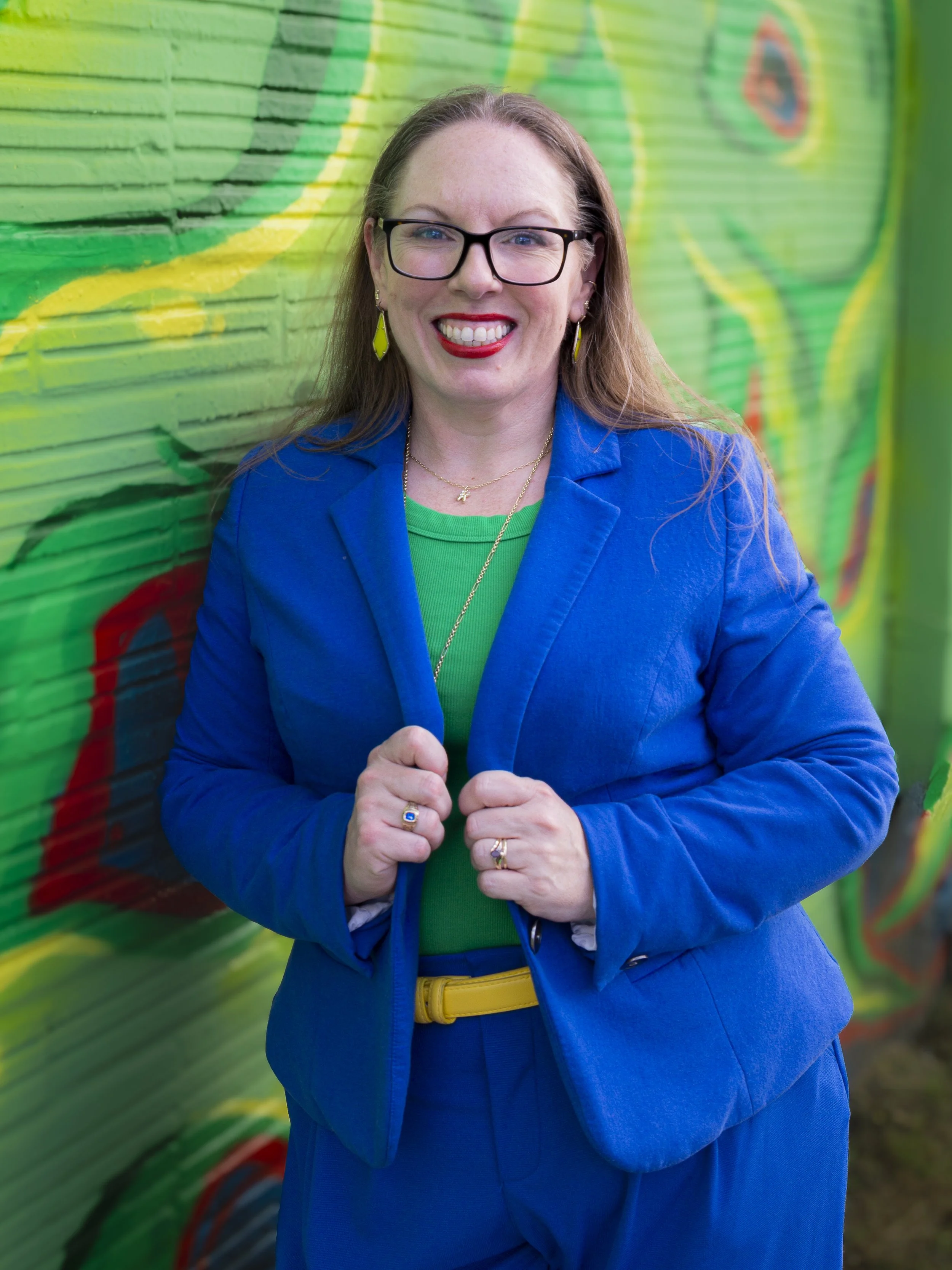 A smiling appellate attorney with long brown hair stands in front of a green graffiti wall. She is wearing a blue blazer and glasses.
