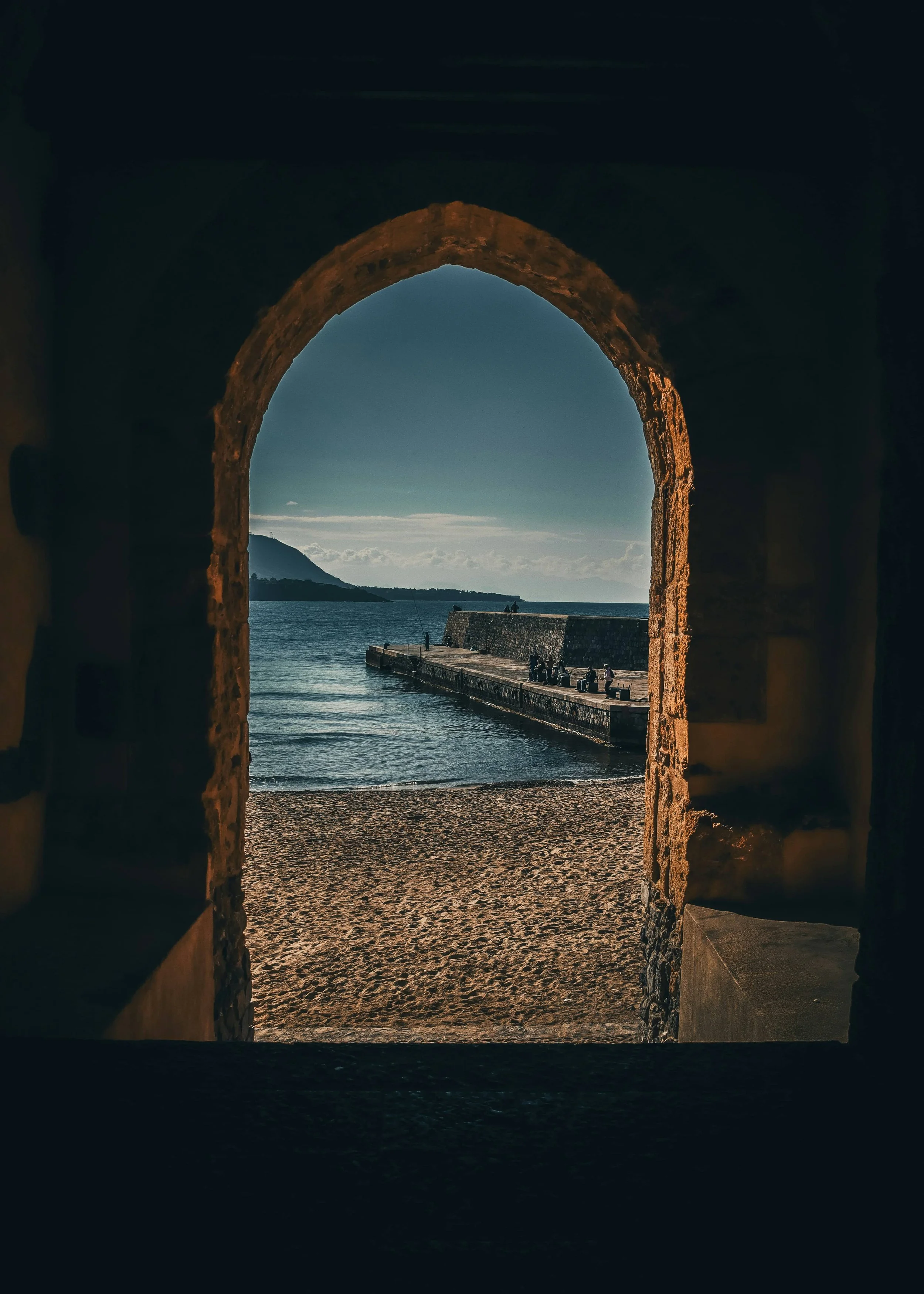 View of a beach scene through a stone archway with a pier extending into the water and a mountain in the background.