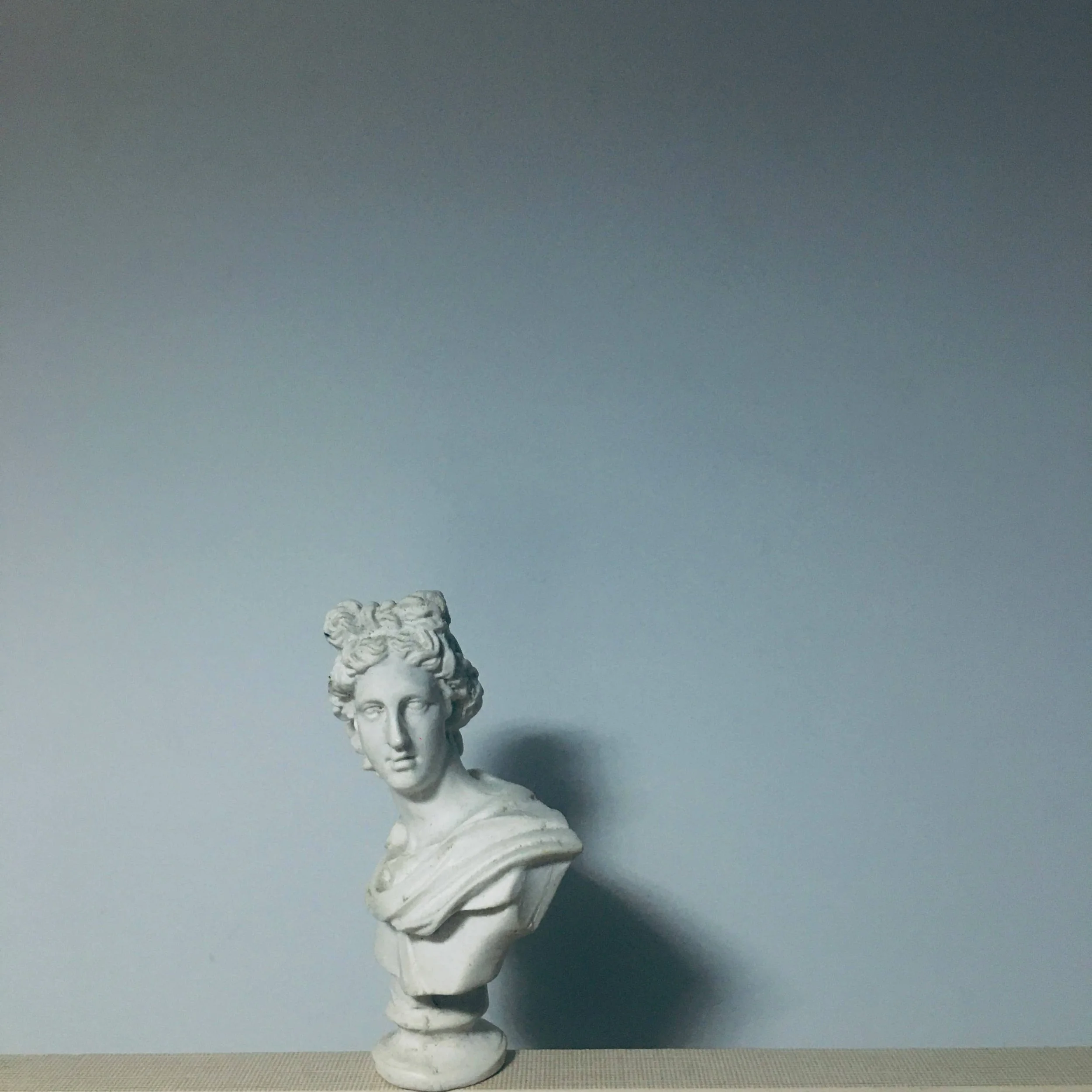 White marble bust sculpture of a woman with styled hair, placed on a beige surface against a plain gray background.