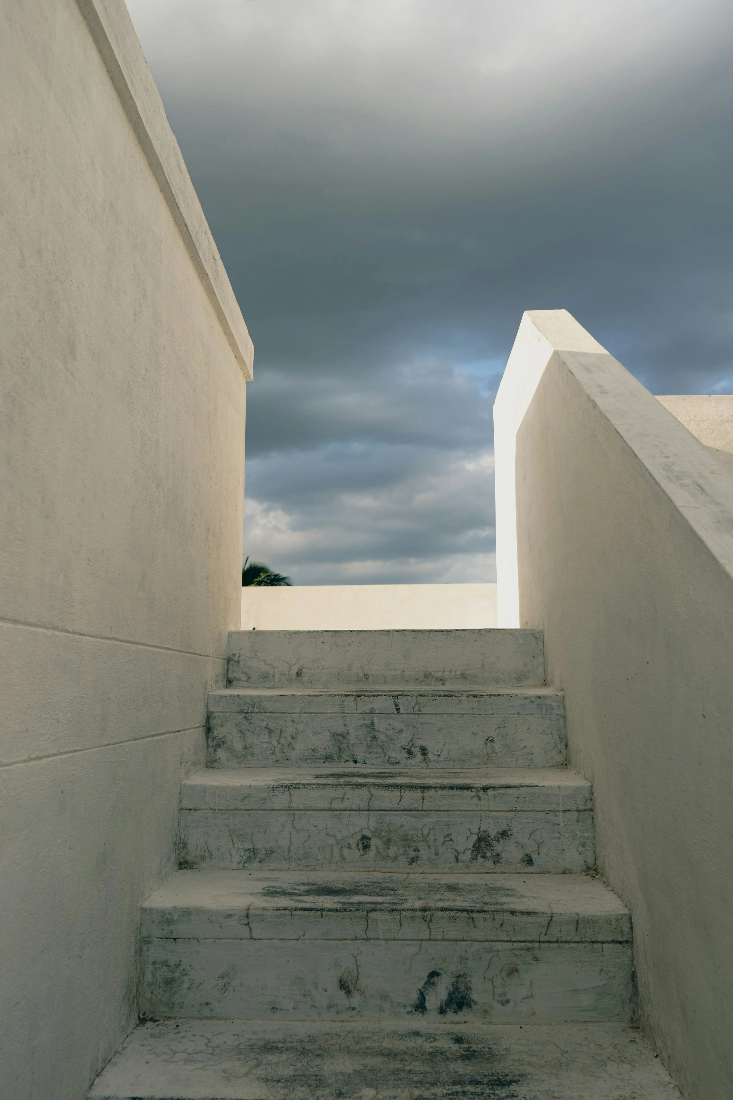 A low-angle view of weathered white concrete stairs and walls, with dark storm clouds in the sky.