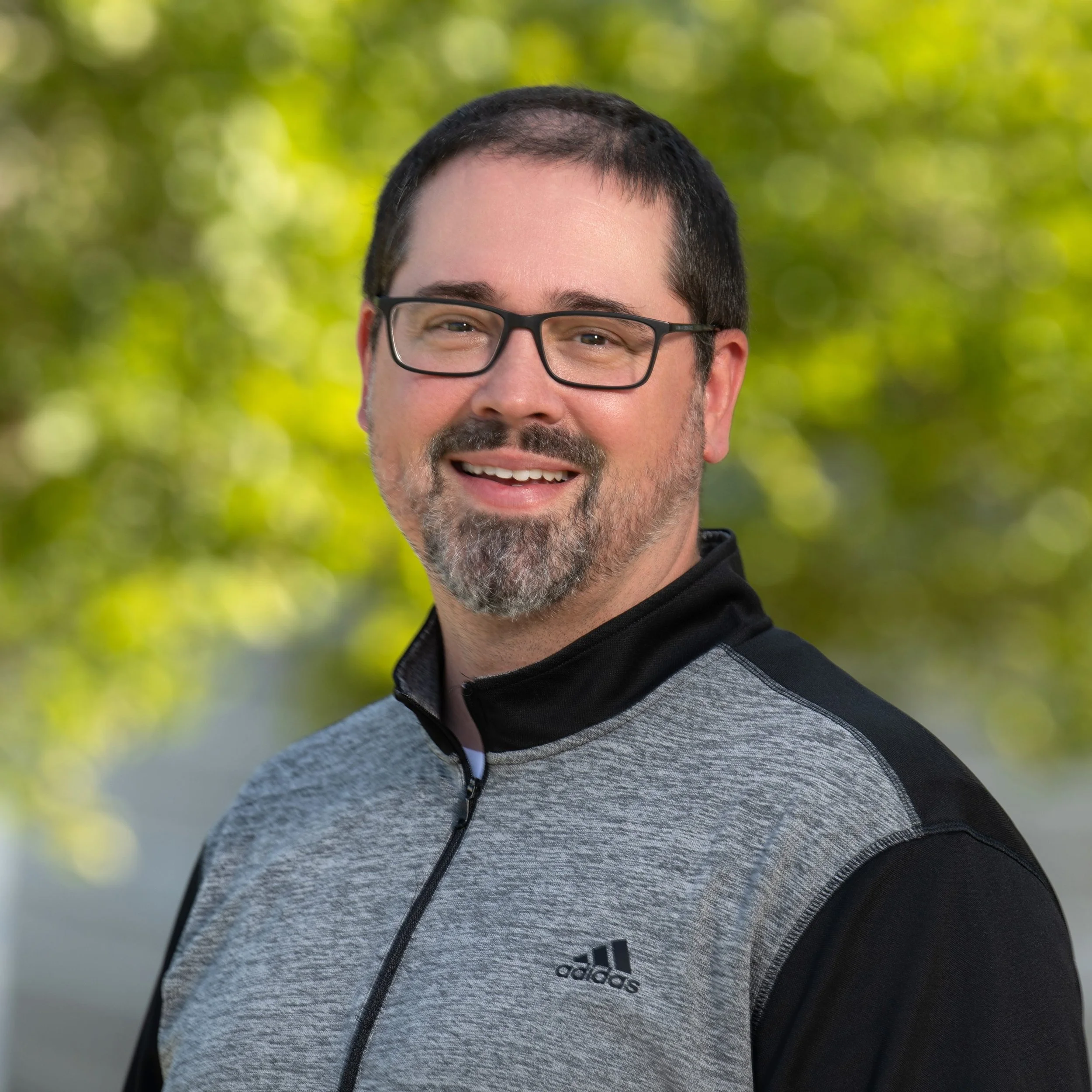A smiling man with dark hair, glasses, and a beard, wearing a gray and black Adidas jacket, standing outdoors with a blurred green and yellow background.