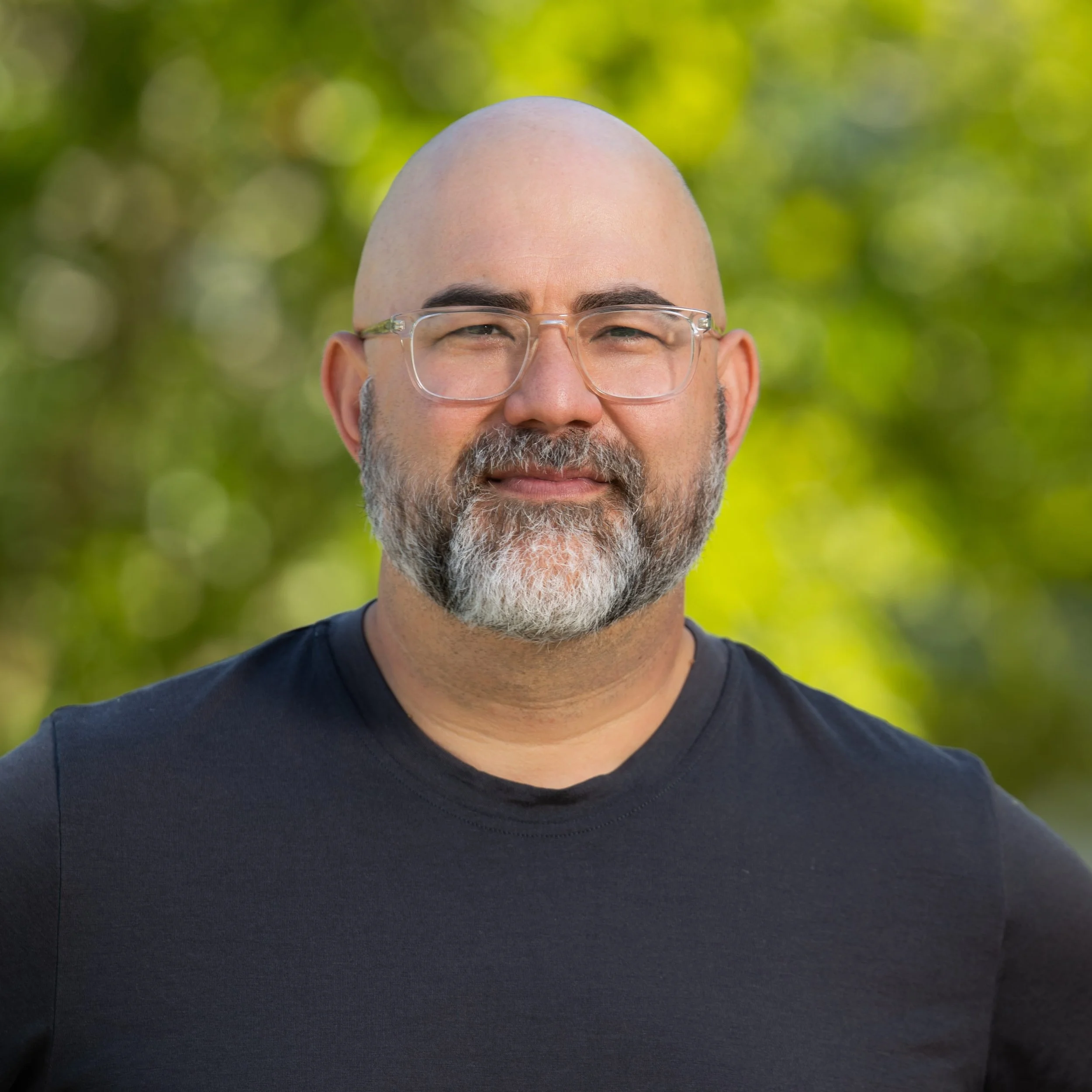 A man with glasses and a beard standing outdoors with a blurred green background.