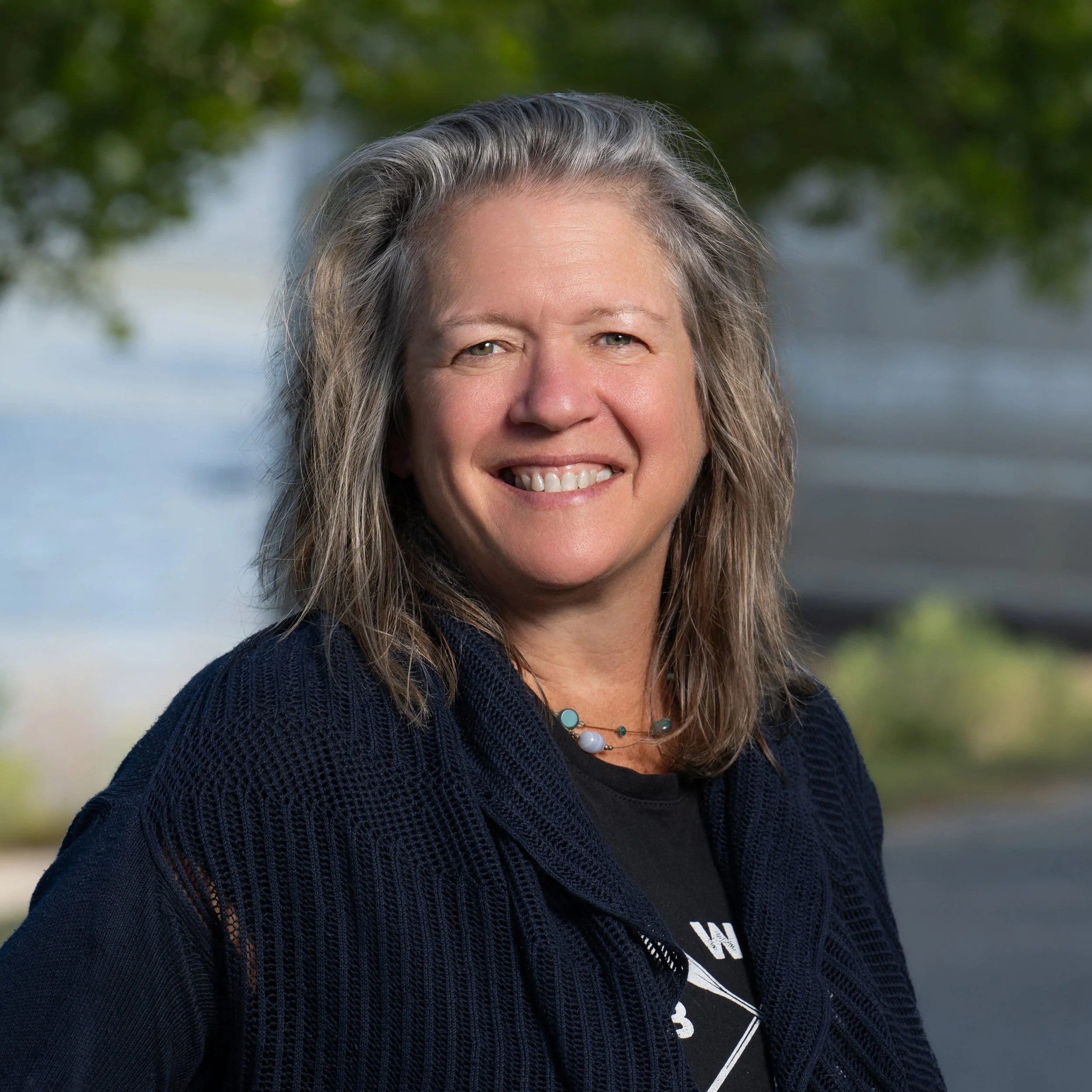 A woman with shoulder-length gray hair smiling outdoors with trees and a body of water in the background.