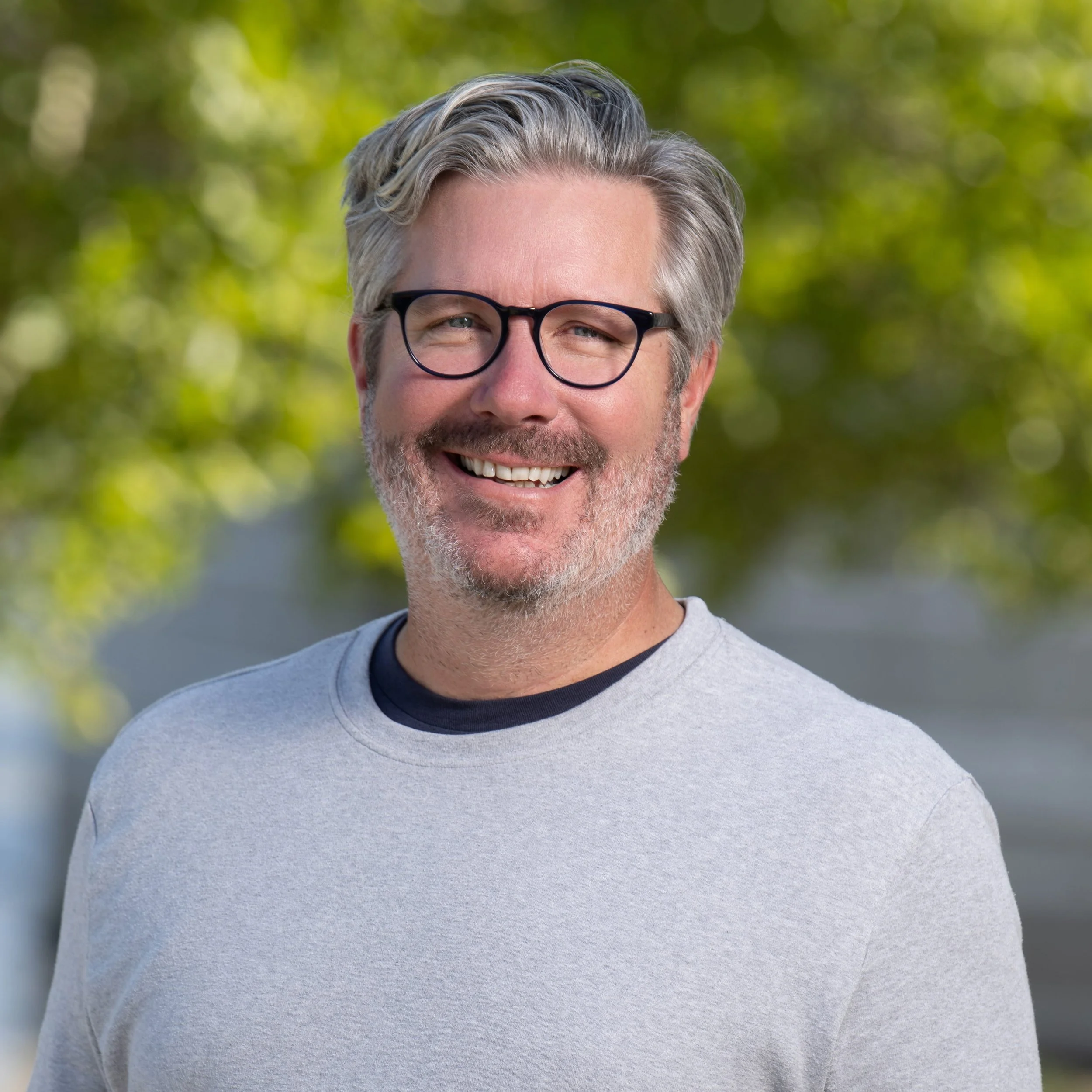 Smiling middle-aged man with glasses, gray hair, and beard, outdoors with blurred green trees in background.