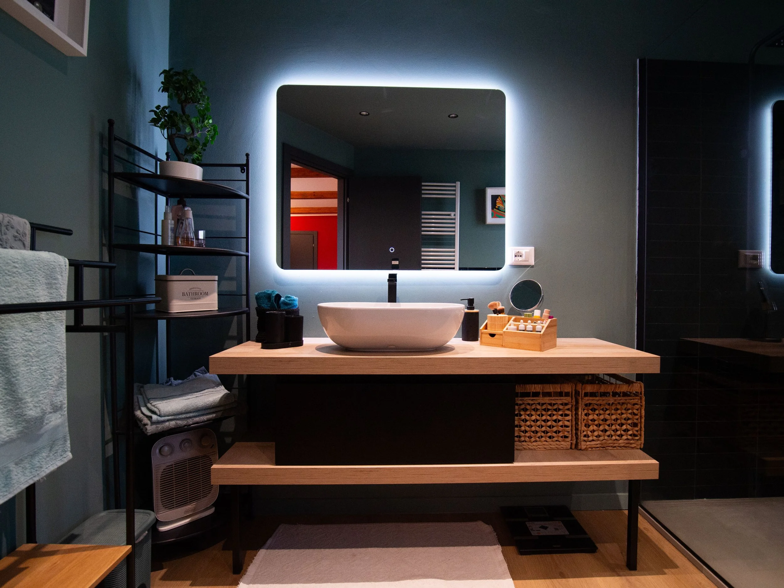 Specchio con retroilluminazione sopra un lavabo bianco su un piano di legno con accessori da bagno, scaffale con piante e asciugamani a sinistra, e parte di una parete di doccia a destra.