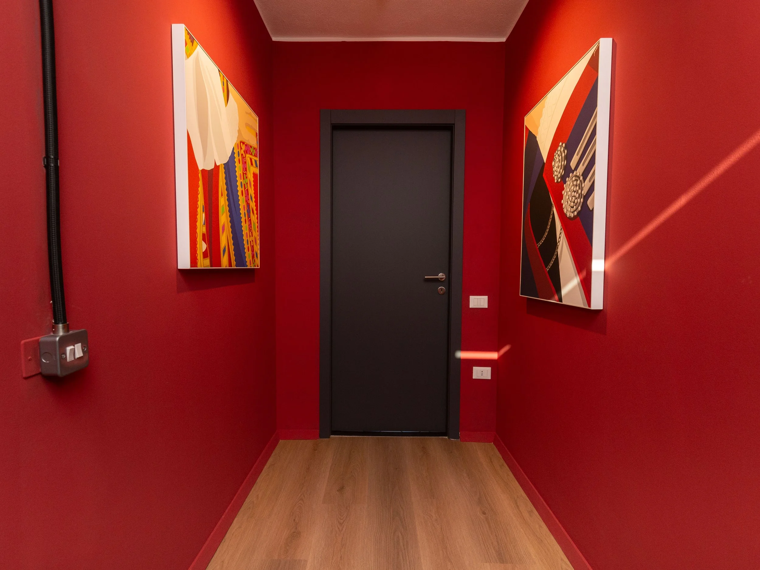 Corridoio con pareti rosse, due quadri astratti appesi, e una porta nera centrale.