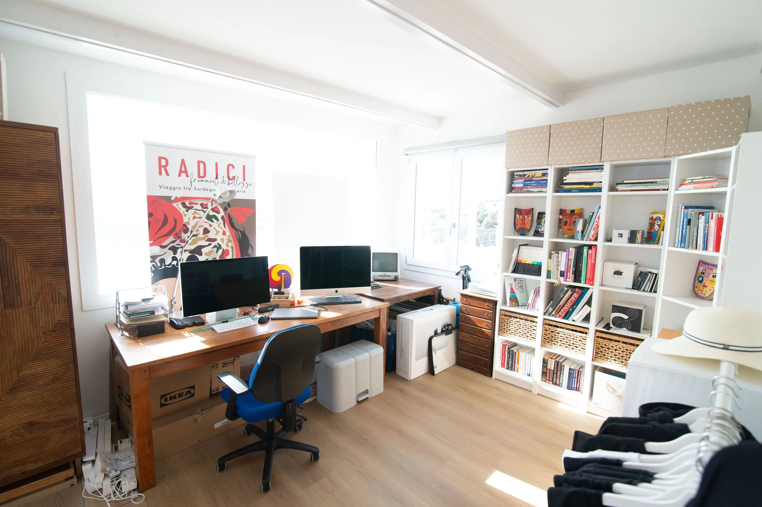 Stanza ordinata con scrivania in legno, due monitor, una sedia da ufficio, scaffali bianchi pieni di libri e decorazioni colorate, finestra che lascia entrare luce naturale, pareti bianche e pavimento in legno chiaro.