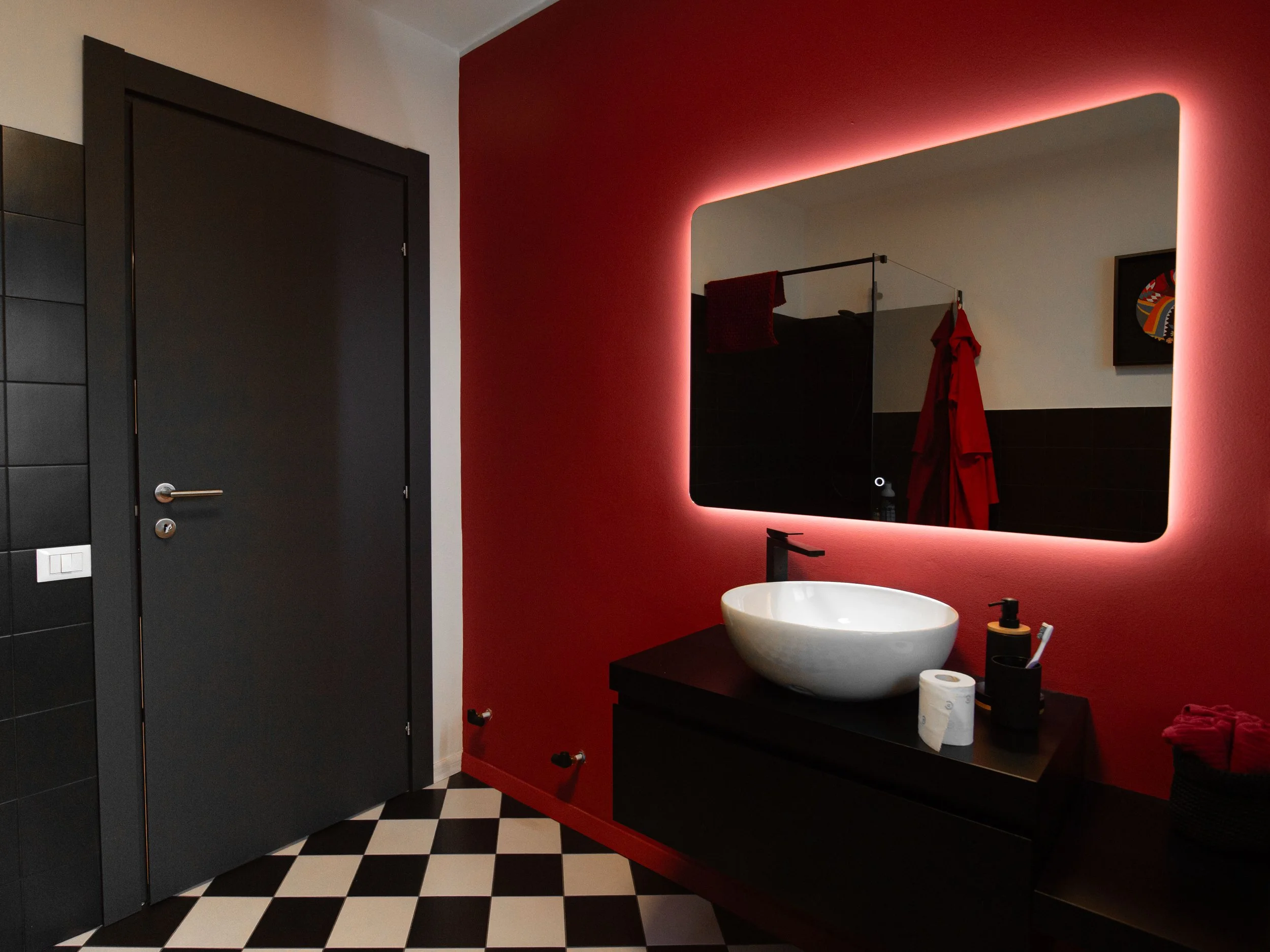 Bagno modernissimo con pareti rosse e dettagli neri, grande specchio con illuminazione a LED, lavabo rotondo bianco su mobile nero, accessori da bagno come dispenser e rotoli di carta, pavimento a scacchi bianco e nero.