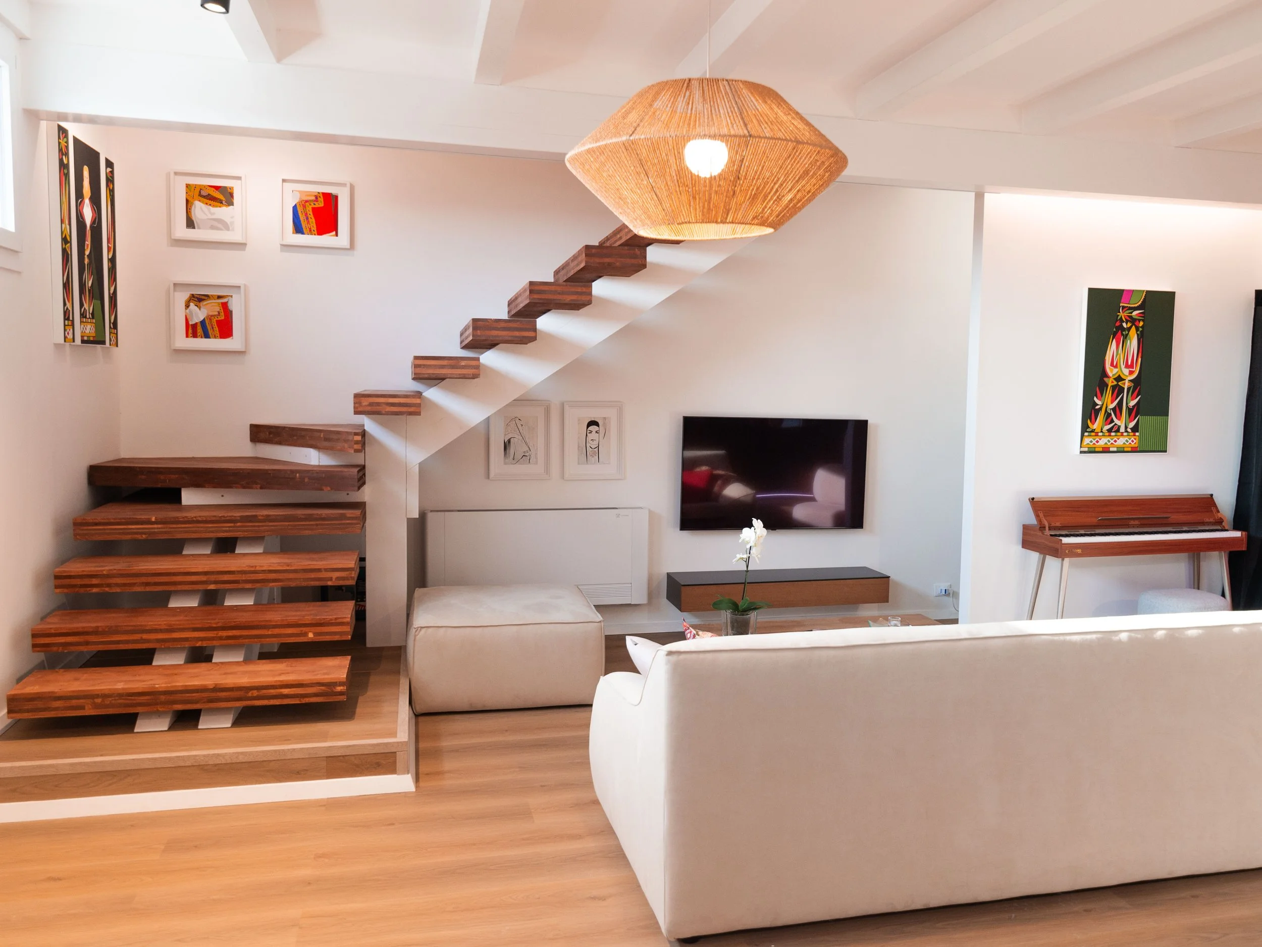 Interno di un soggiorno con divano bianco, televisore a parete, quadro colorato e scala in legno che conduce a un piano superiore.