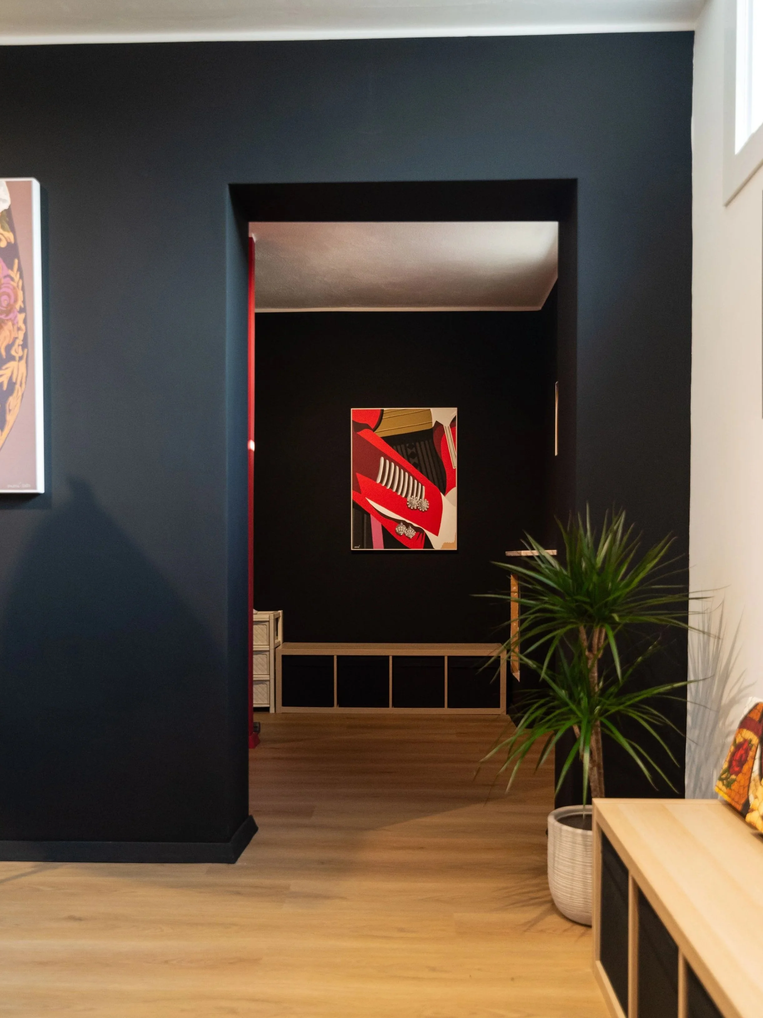 Interno di una stanza con pareti nere, un quadro con forme geometriche e colori vivaci, e un grande vaso con pianta verde in primo piano.