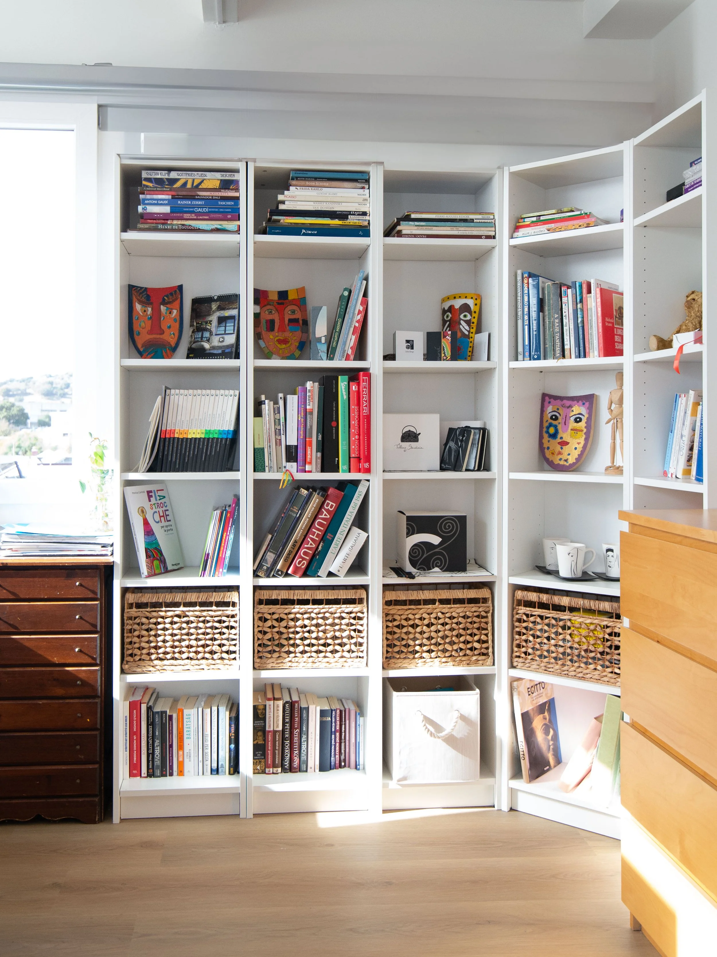 Scaffale bianco con libri, lavori artistici e decorazioni, tra cui maschere colorate, e cesti di vimini, in una stanza luminosa con parquet.