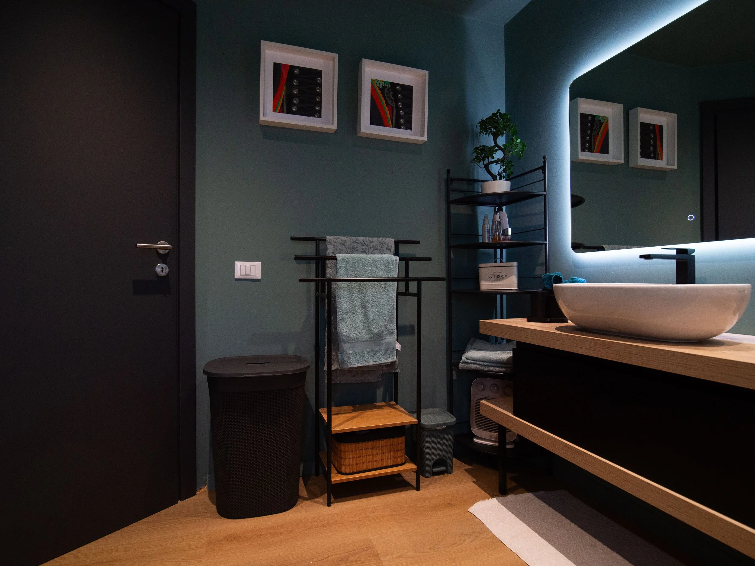 Bagno moderno con lavabo in piedi, specchio con illuminazione a LED, scaffale nero con piante e prodotti, asciugamani appesi, lavatrice, cesto per la biancheria, pareti verdi e pavimento in legno.