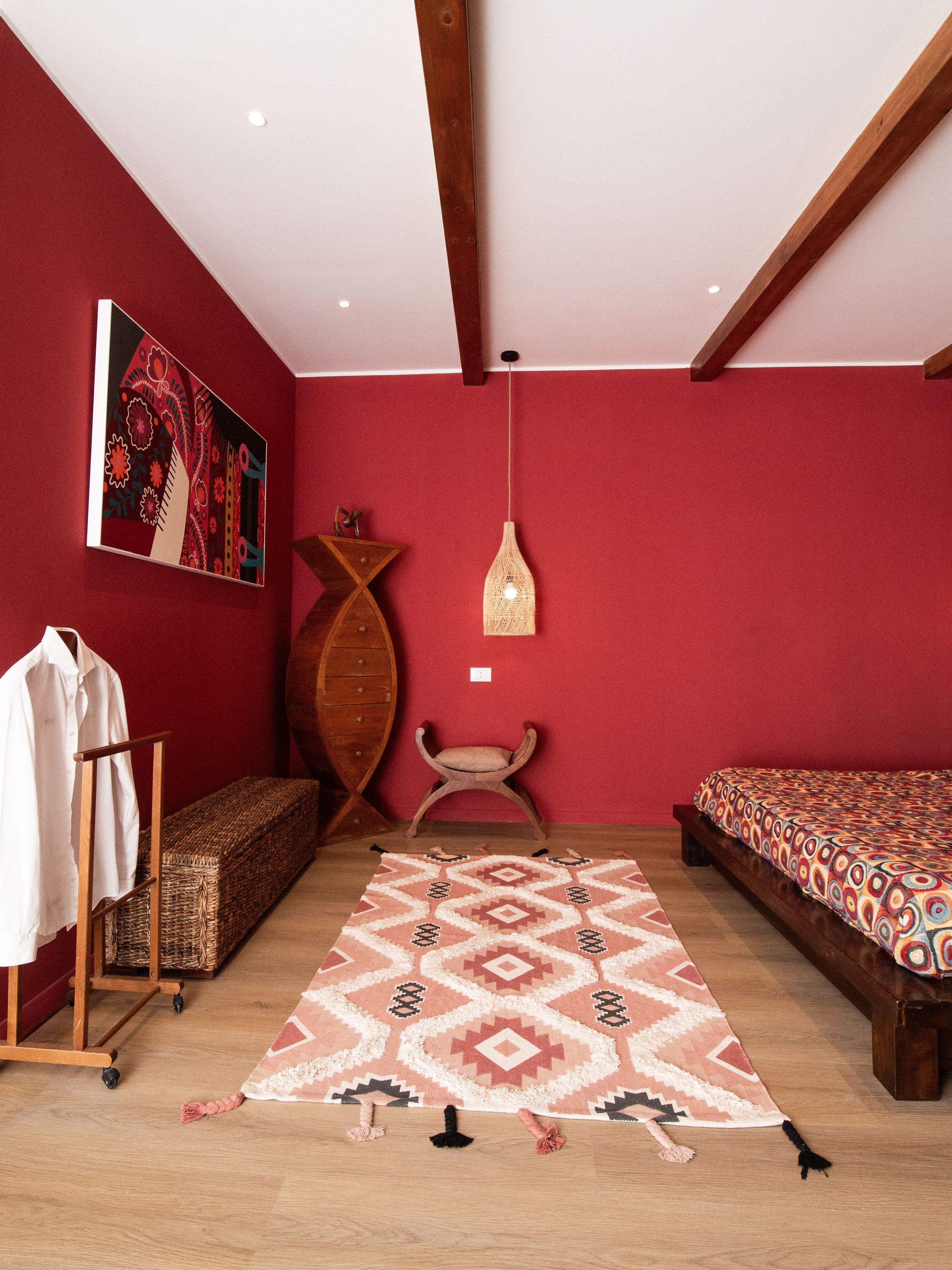 Stanza con pareti rosse, letto con copriletto colorato, tappeto con motivi geometrici, lampada di vimini,seduta in stile antichizzato, armadio abiti e quadro con pattern colorato.