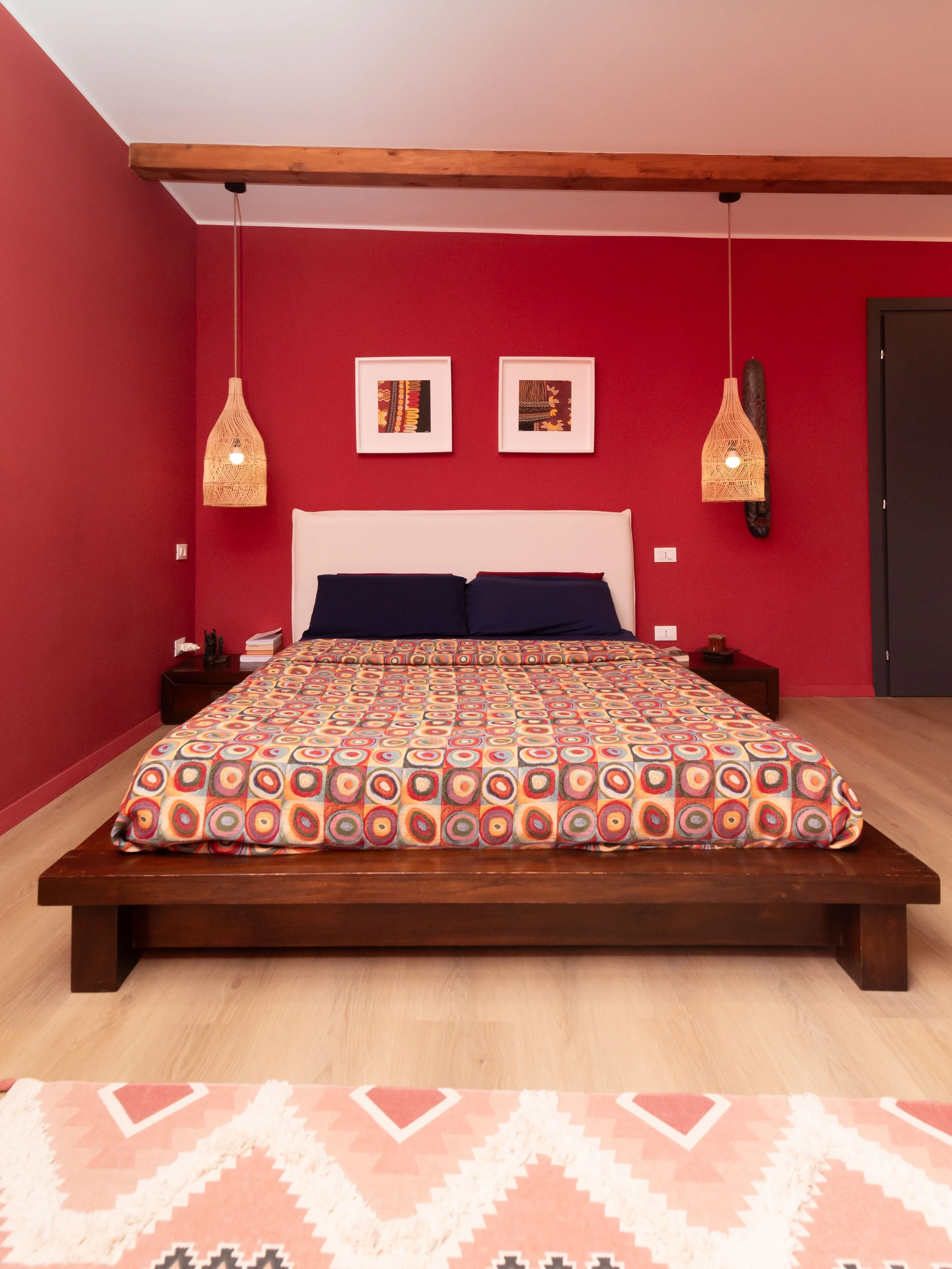 Camera da letto con parete rossa, letto con testiera bianca, copriletto con motivo a cerchi colorati, due quadri astratti sopra il letto, due lampade a sospensione con paralumi di vimini ai lati del letto.