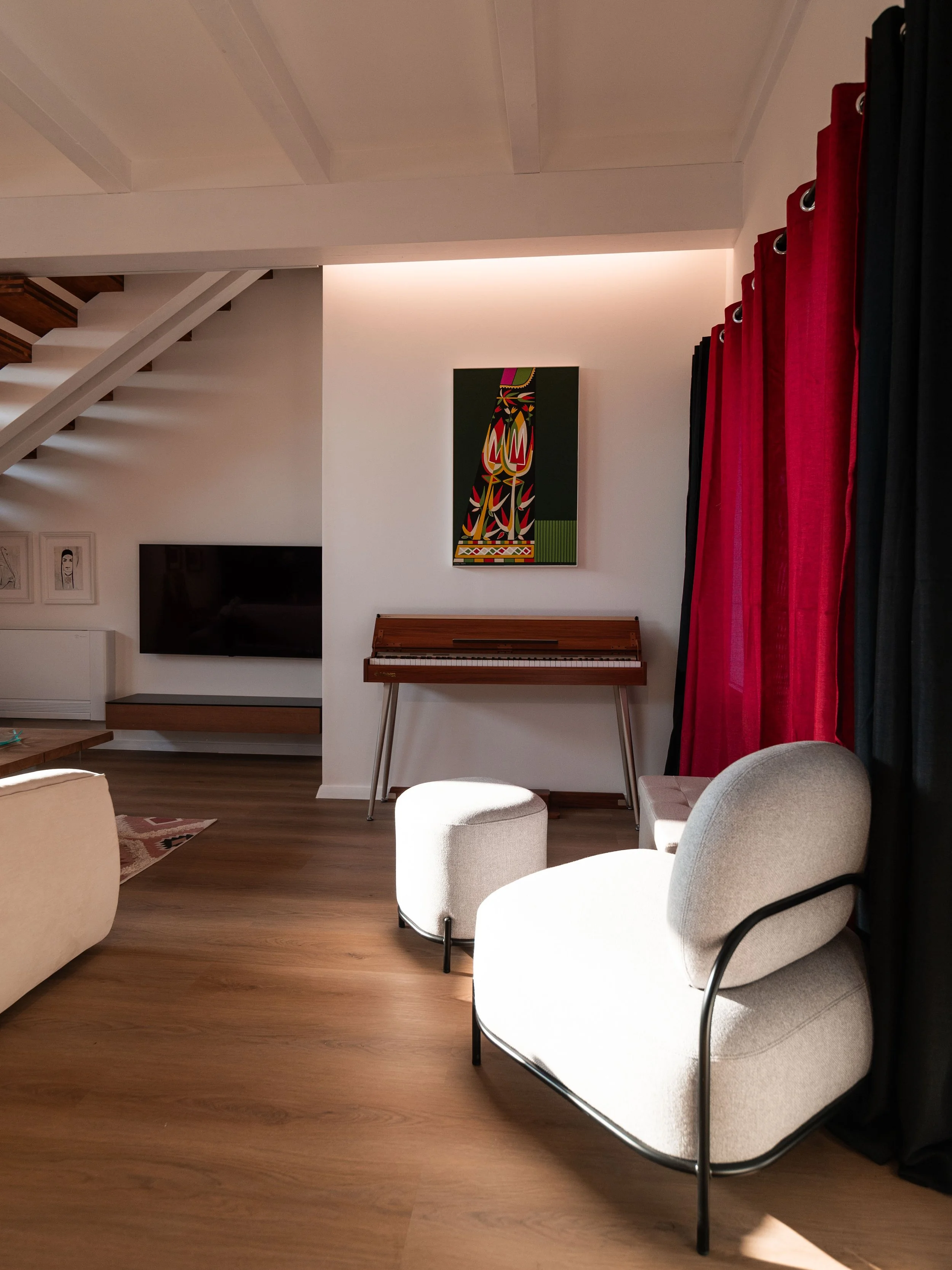 Soggiorno con poltrona beige, pouf, tende rosse e nere, dipinto colorato e pianoforte in legno.