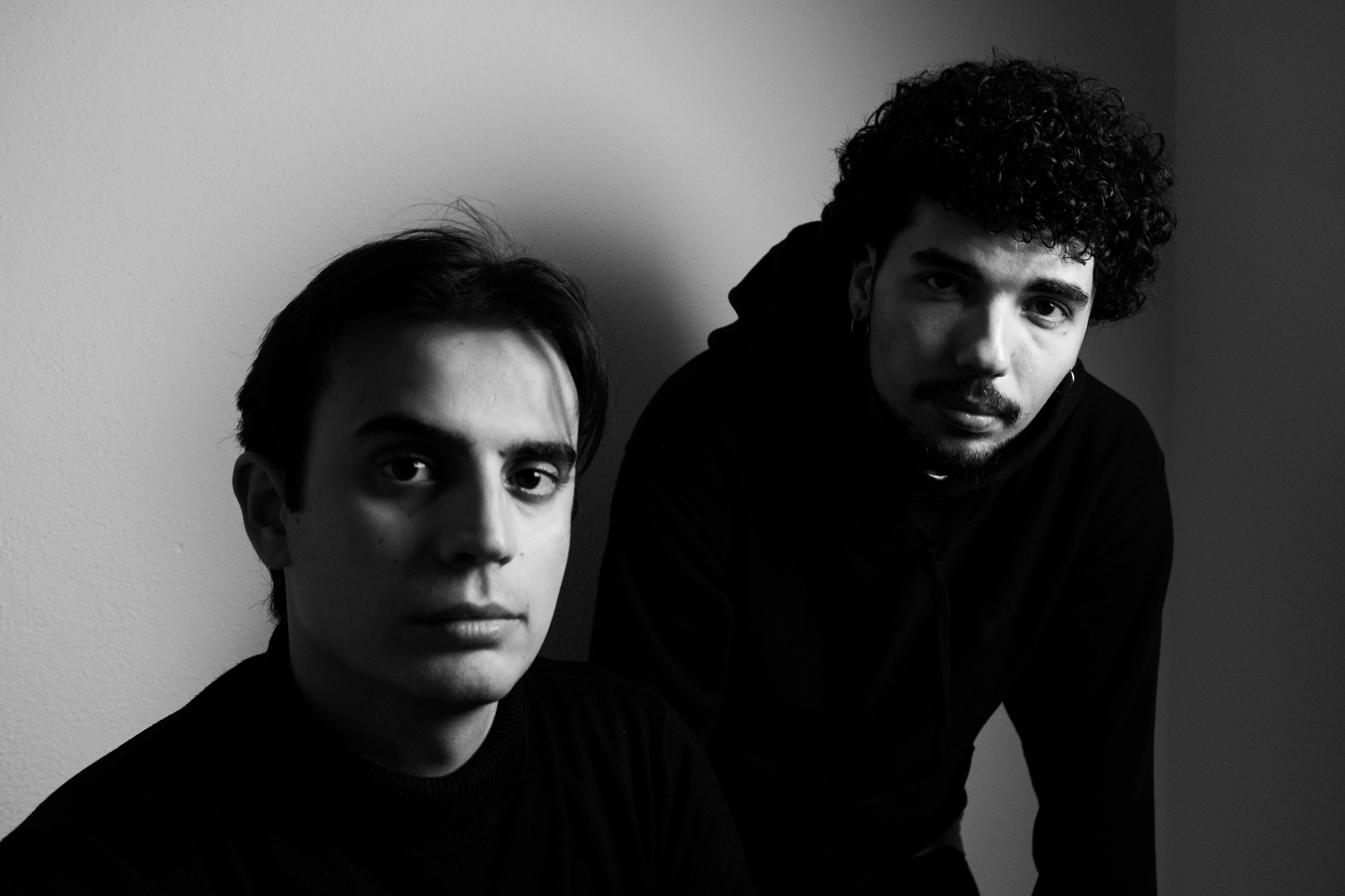 Due uomini con capelli scuri, uno con capelli corti e l'altro con capelli ricci, seduti contro una parete bianca, in foto in bianco e nero.