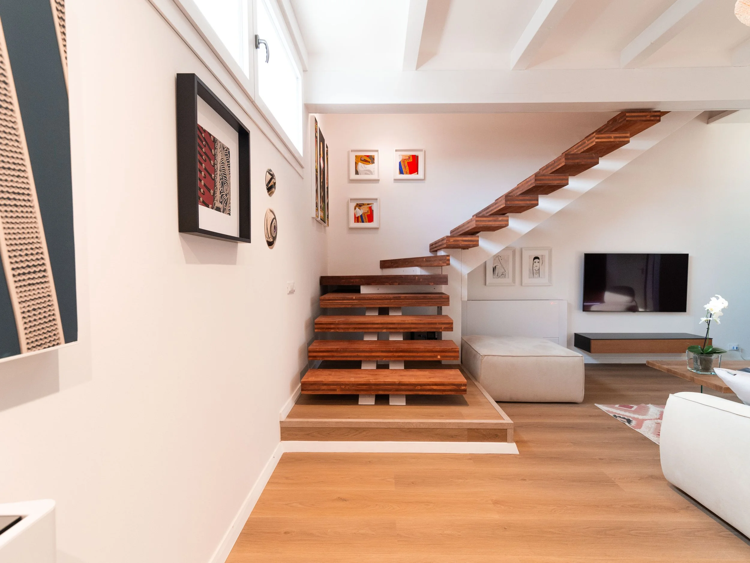 Interno moderno con pareti bianche, arredi minimalisti, un divano beige, un televisore a parete, un tavolino con una pianta e un'installazione di quadri e decorazioni sulla parete, e una scala di legno che sale verso un soppalco superiore.