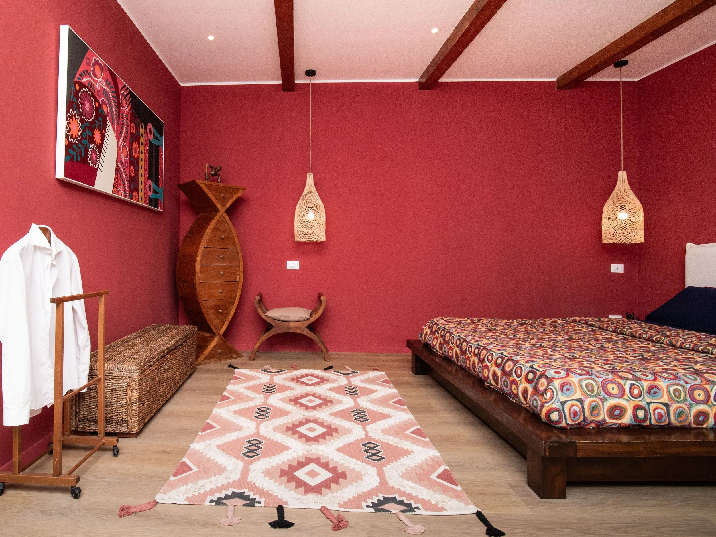 Camera da letto con pareti color terracotta, letto con testiera bianca e piumone con motivo circolare colorato, tappeto con motivo geometrico rosa, bianco e nero, lampade di vimini, arredi in legno e un quadro astratto con colori vivaci.