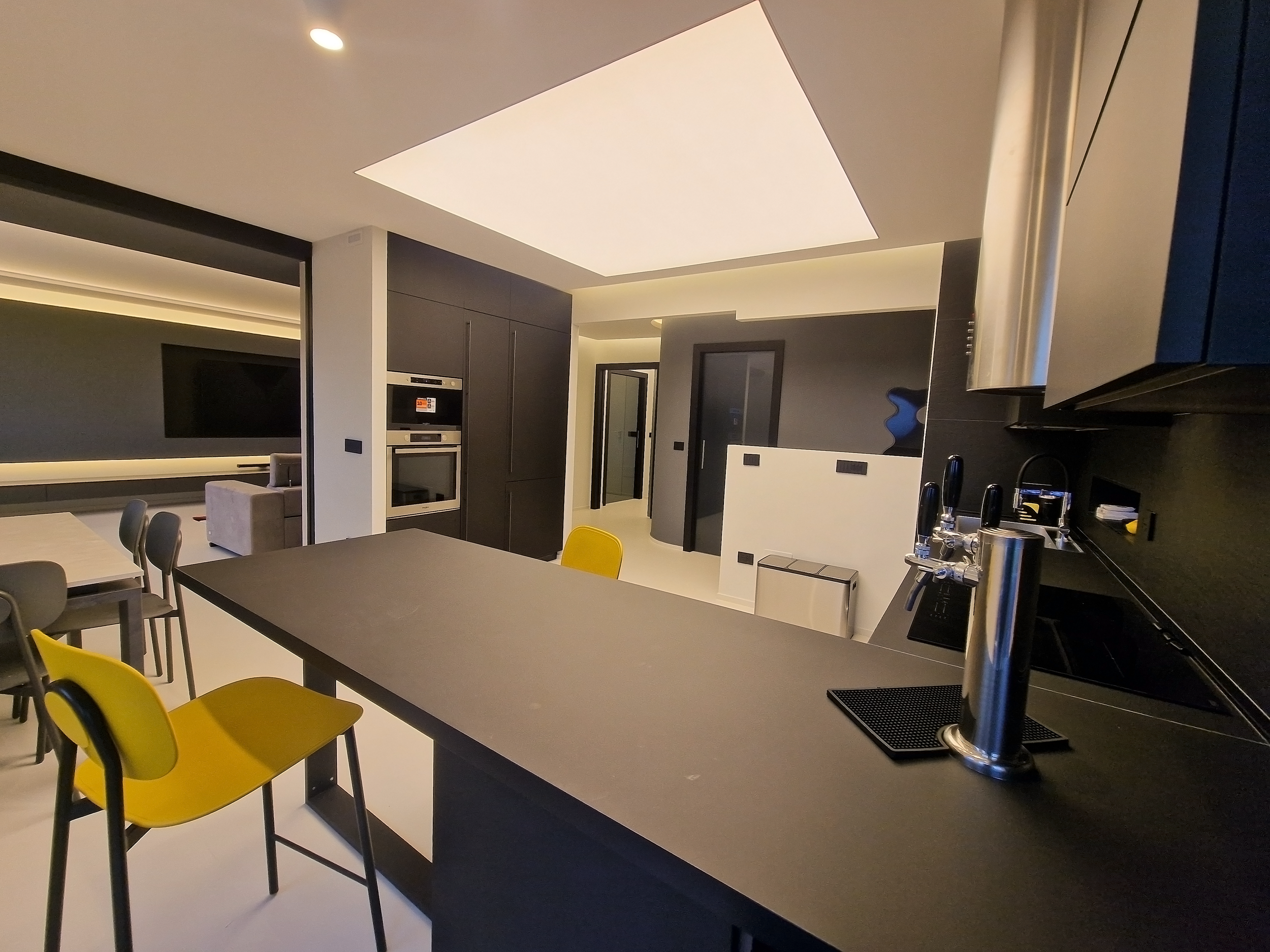 Interno di una cucina moderna con piano di lavoro nero, sedie gialle e nere, con soggiorno visibile sullo sfondo, e pareti scure.