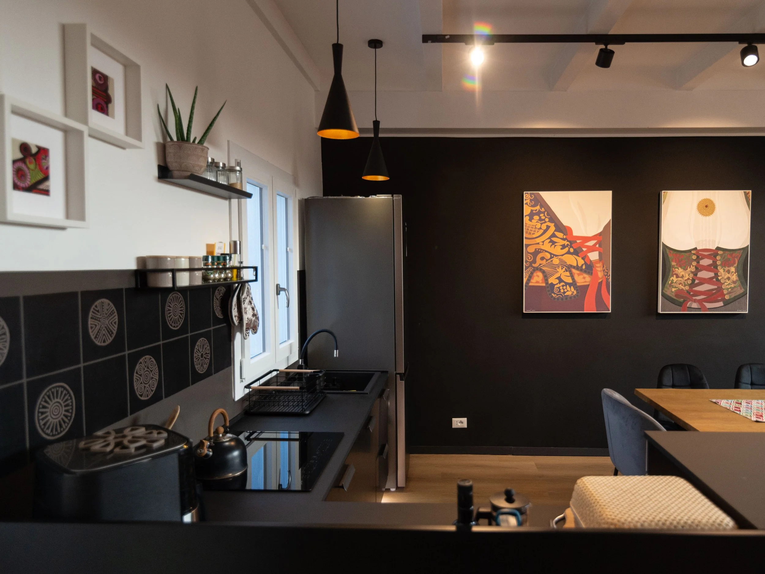 Interior di una cucina moderna con pareti nere, decorazioni minimaliste, elettrodomestici in acciaio inox, piantina in vaso e quadri artistici appesi alla parete.