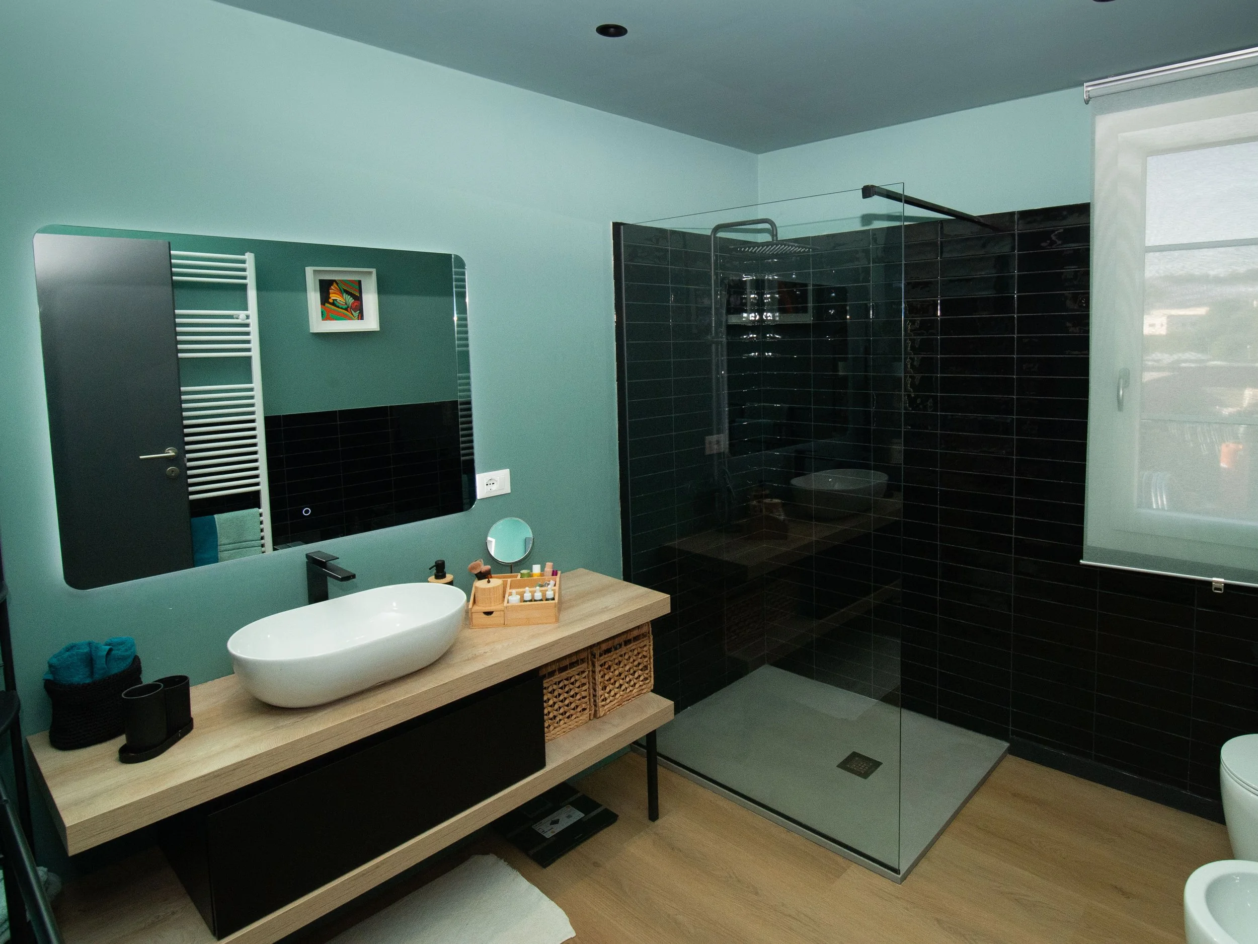 Bagno moderno con pareti verdi e parete di doccia con piastrelle nere, grande specchio sopra il lavabo, lavabo a bacinella bianco su mobile in legno con cassetti, asciugamani e vari prodotti di bellezza sugli scaffali.