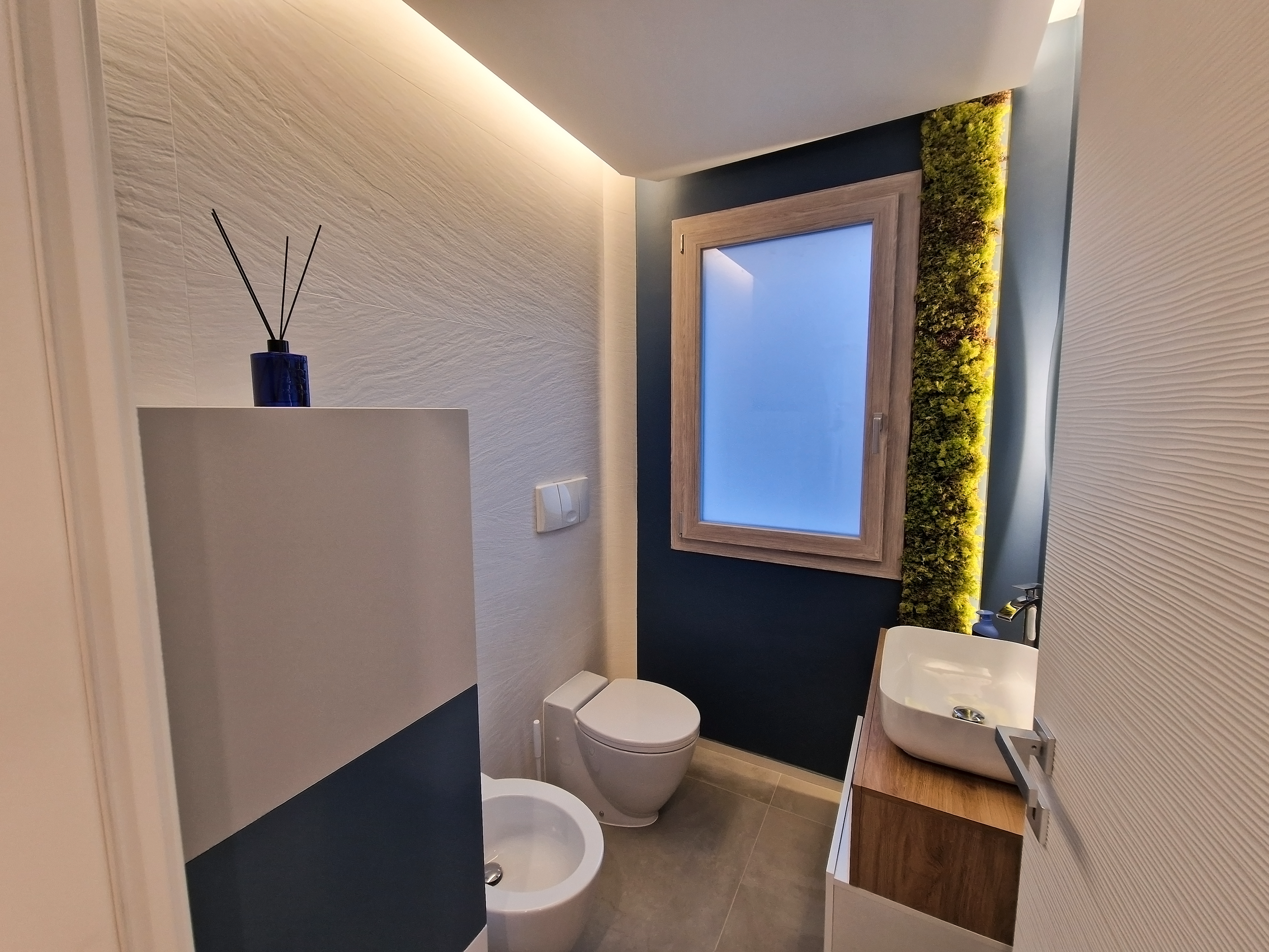 Bagno con WC, bidet, lavabo su mobile in legno, finestra grande con cornice in legno e parete con tessuto botanico verde.