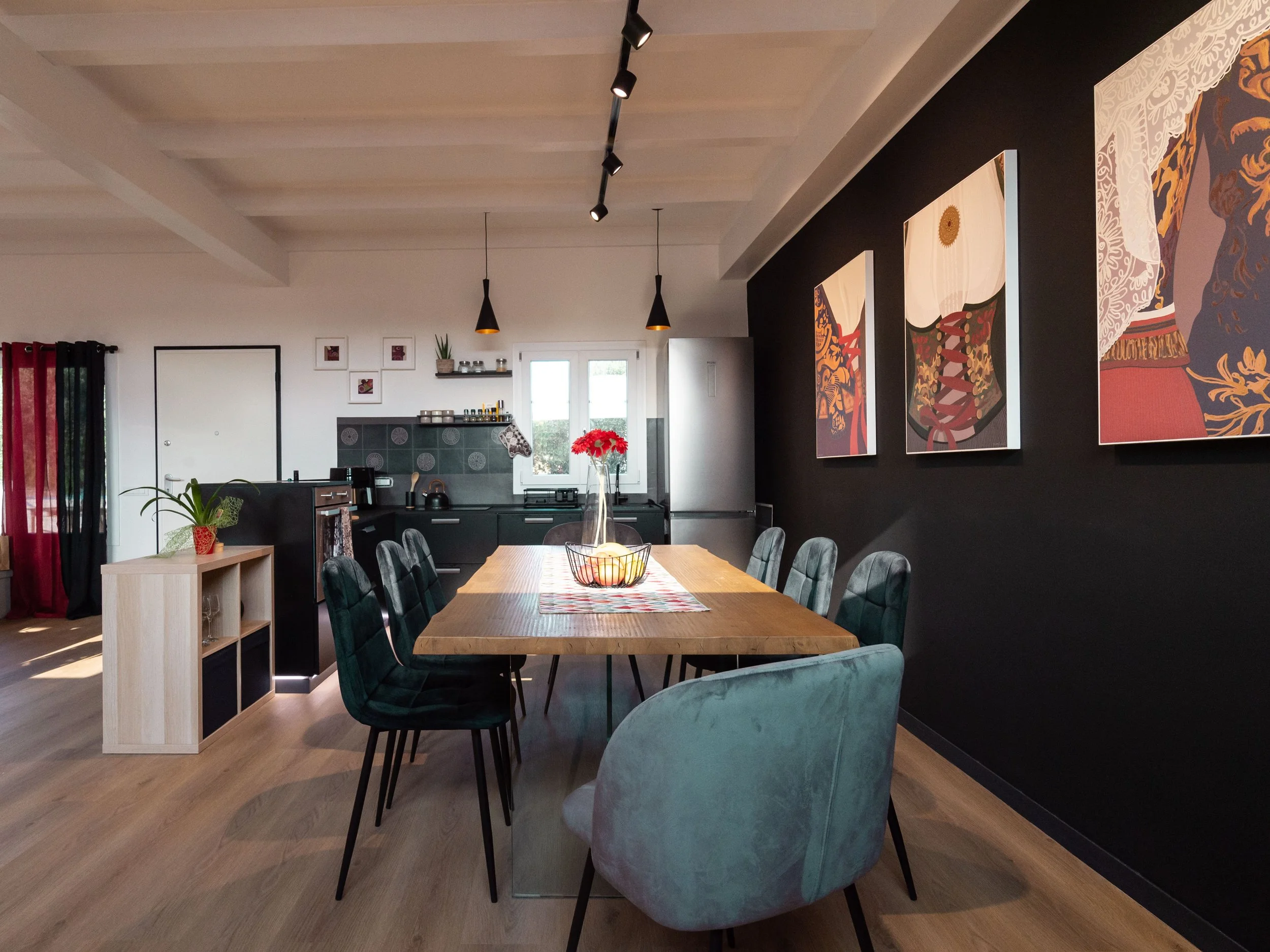 Soggiorno con tavolo da pranzo in legno circondato da sedie, pareti nere con quadri colorati, cucina moderna sullo sfondo con pensili neri, tavolo decorato con un vaso di fiori rossi.