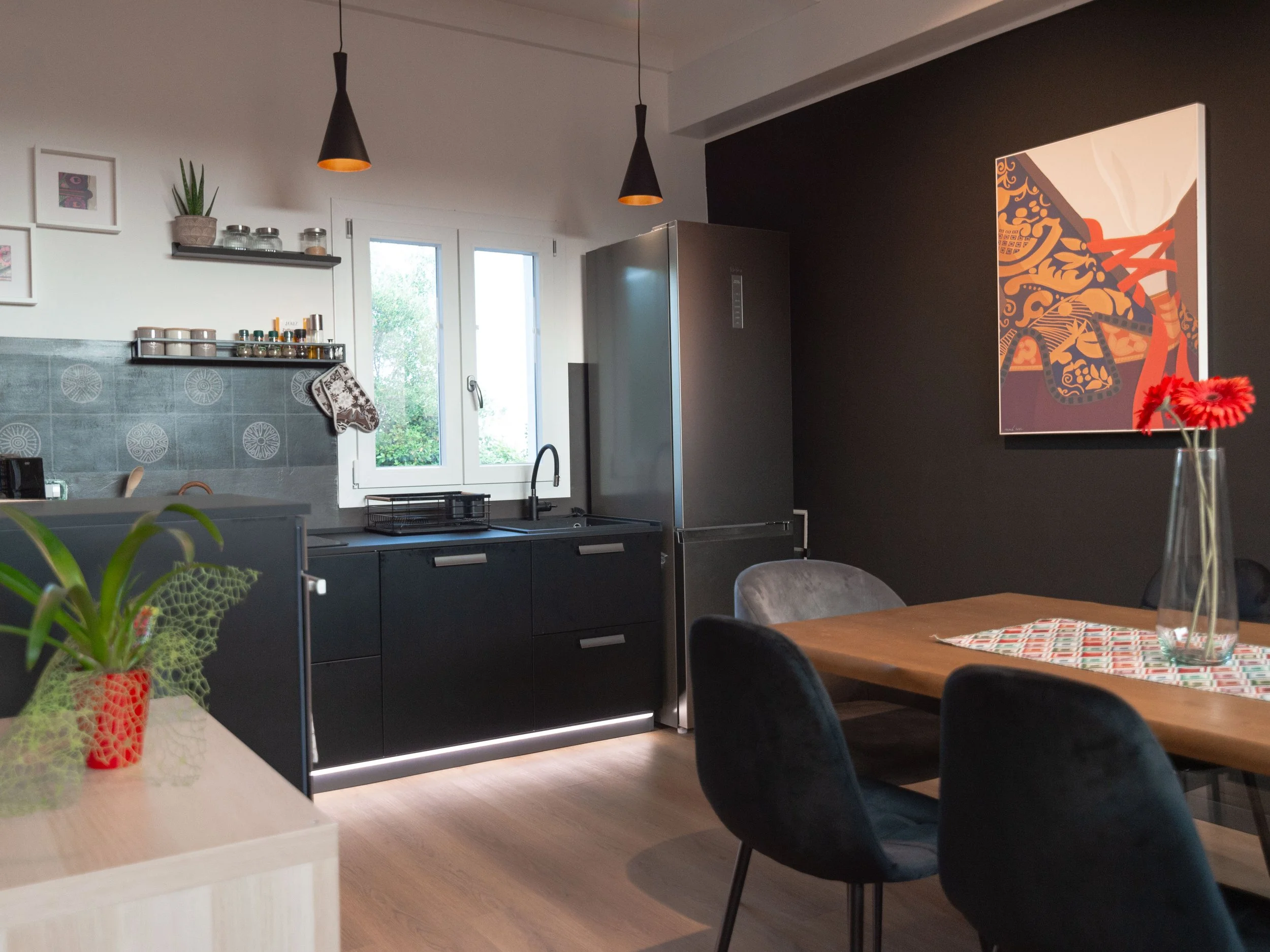 Interno di una cucina moderna con pareti bianche e nere, tavolo in legno con sedie nere, elettrodomestici in acciaio, lampade nere a soffitto, quadri e piante decorative.