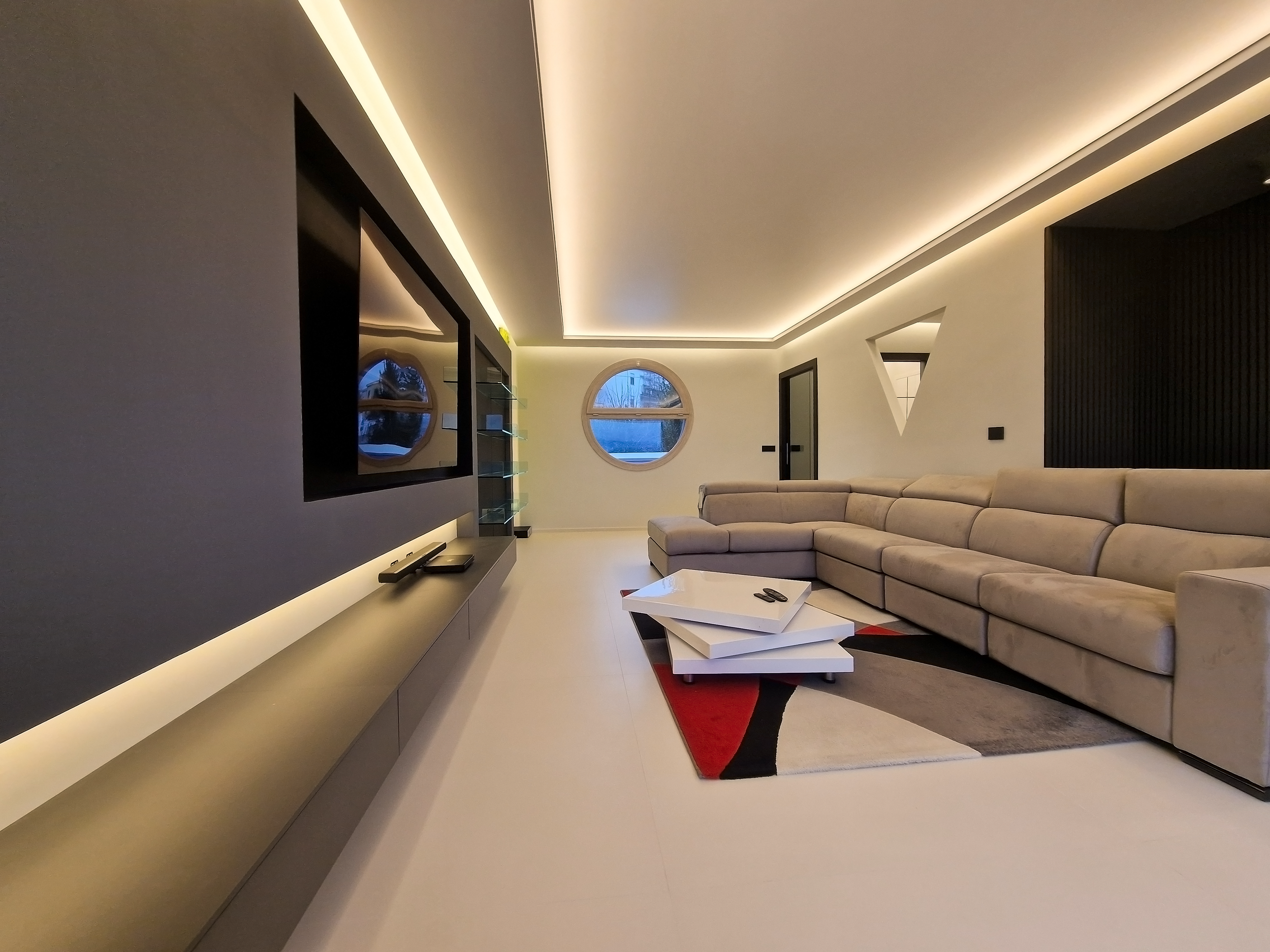 Interno di un soggiorno moderno con grande divano beige, tavolino bianco, pareti illuminate e finestre rotonde.