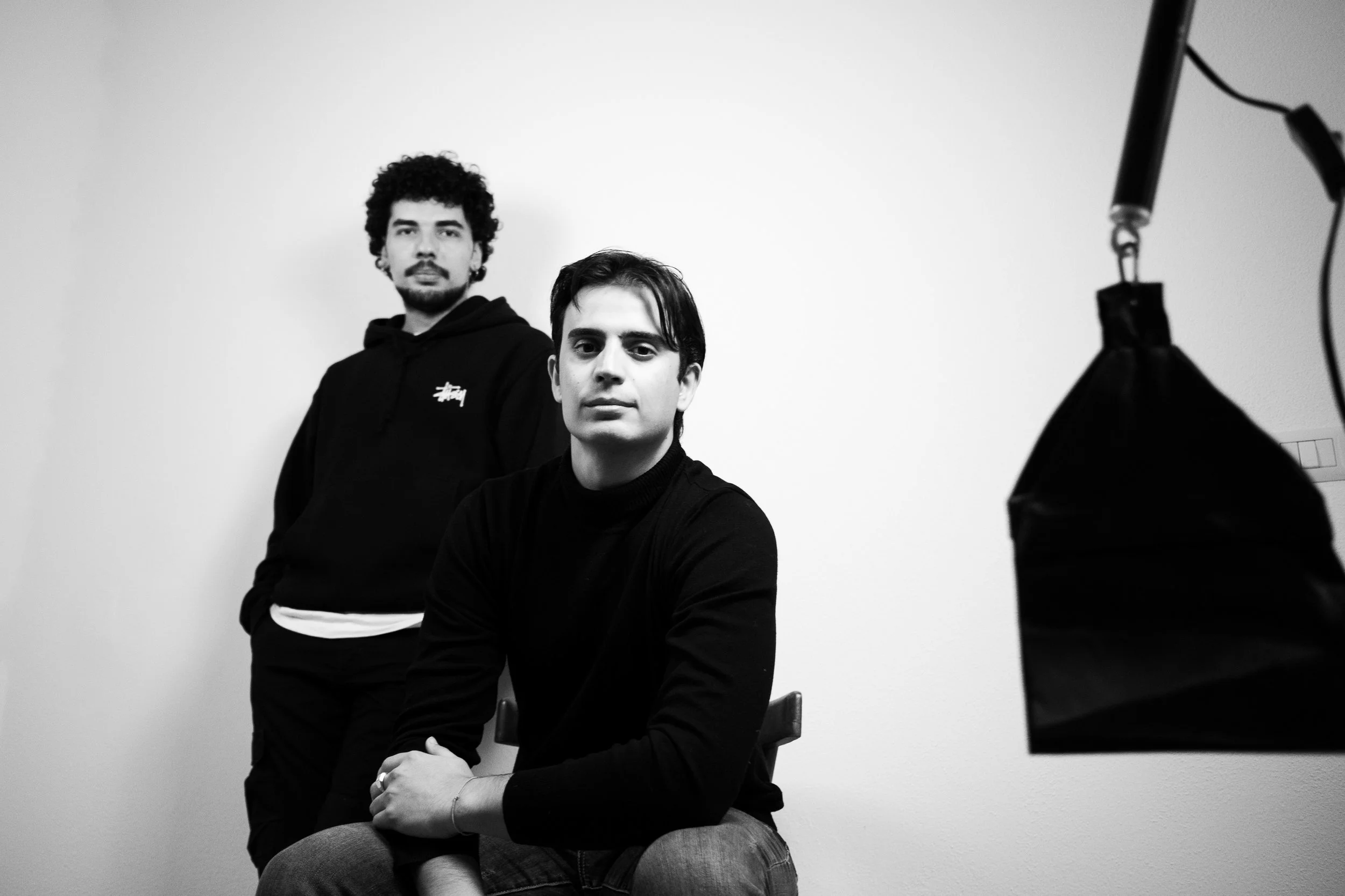 Due giovani uomini in uno studio fotografico, uno seduto e l'altro in piedi, entrambi con espressioni serie, sfondo bianco, illuminazione artificiale visibile a destra.