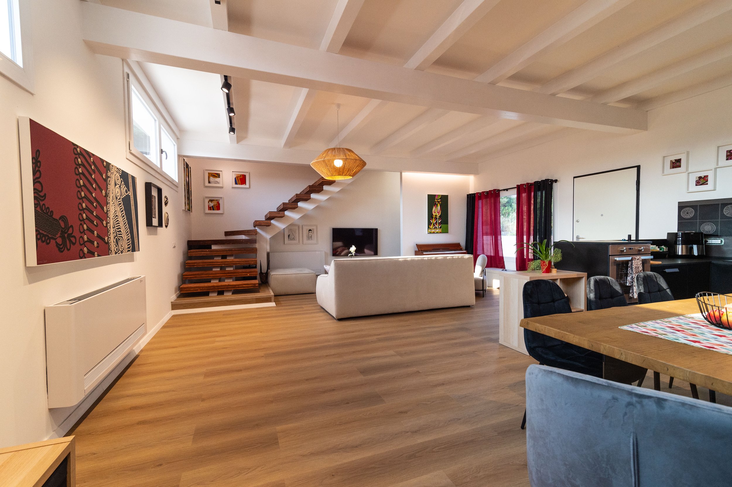 Soggiorno aperto con divano bianco, zona pranzo con tavolo e sedie, cucina in nero, pareti decorate con quadri e arazzi, grandi finestre con tende rosse e nere, lampada di carta sospesa, scala di legno che porta ai piani superiori.