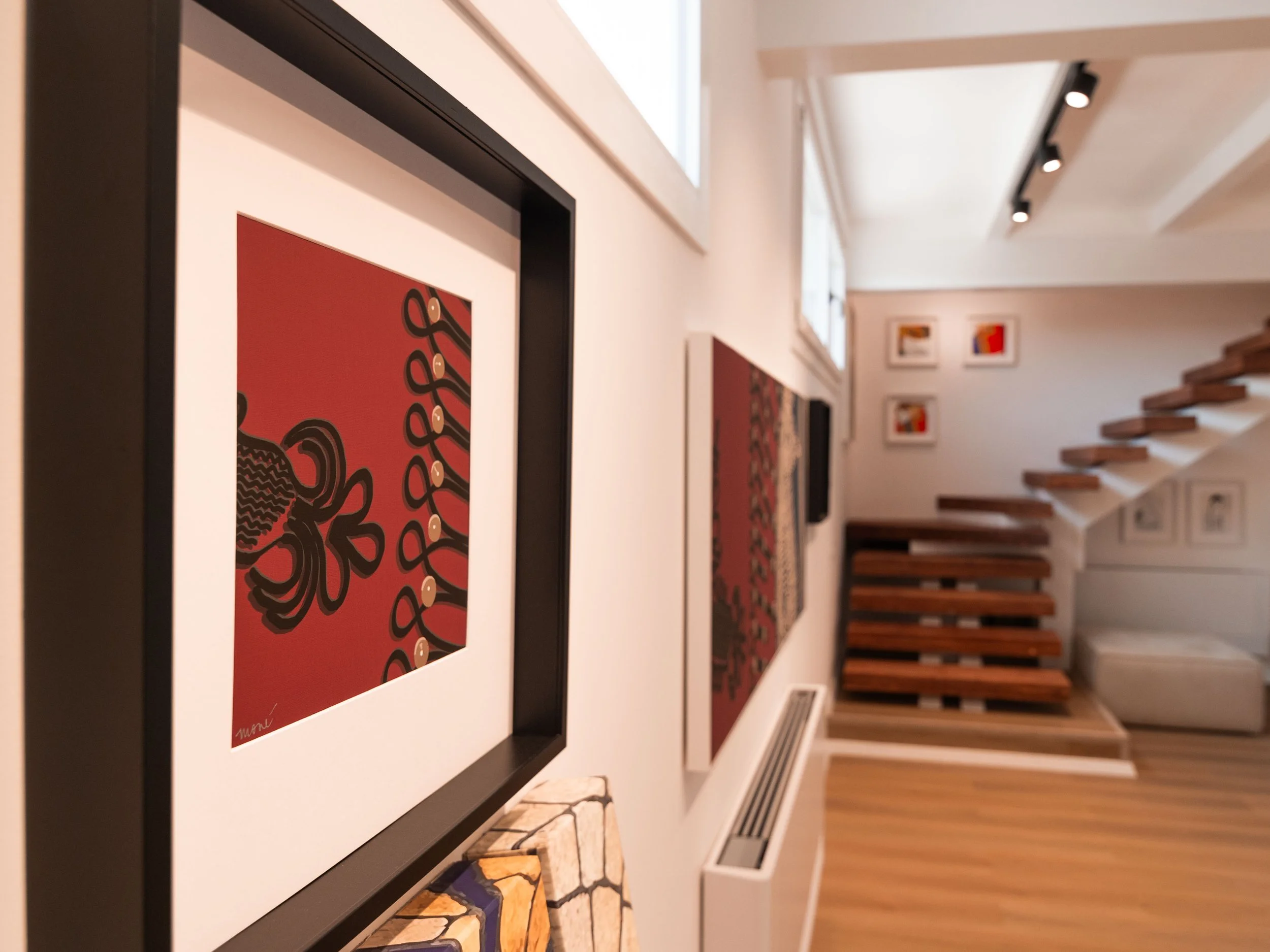 Una parete bianca con diverse opere d'arte appese, tra cui un quadro con sfondo rosso e disegni neri, e una scalinata in legno.