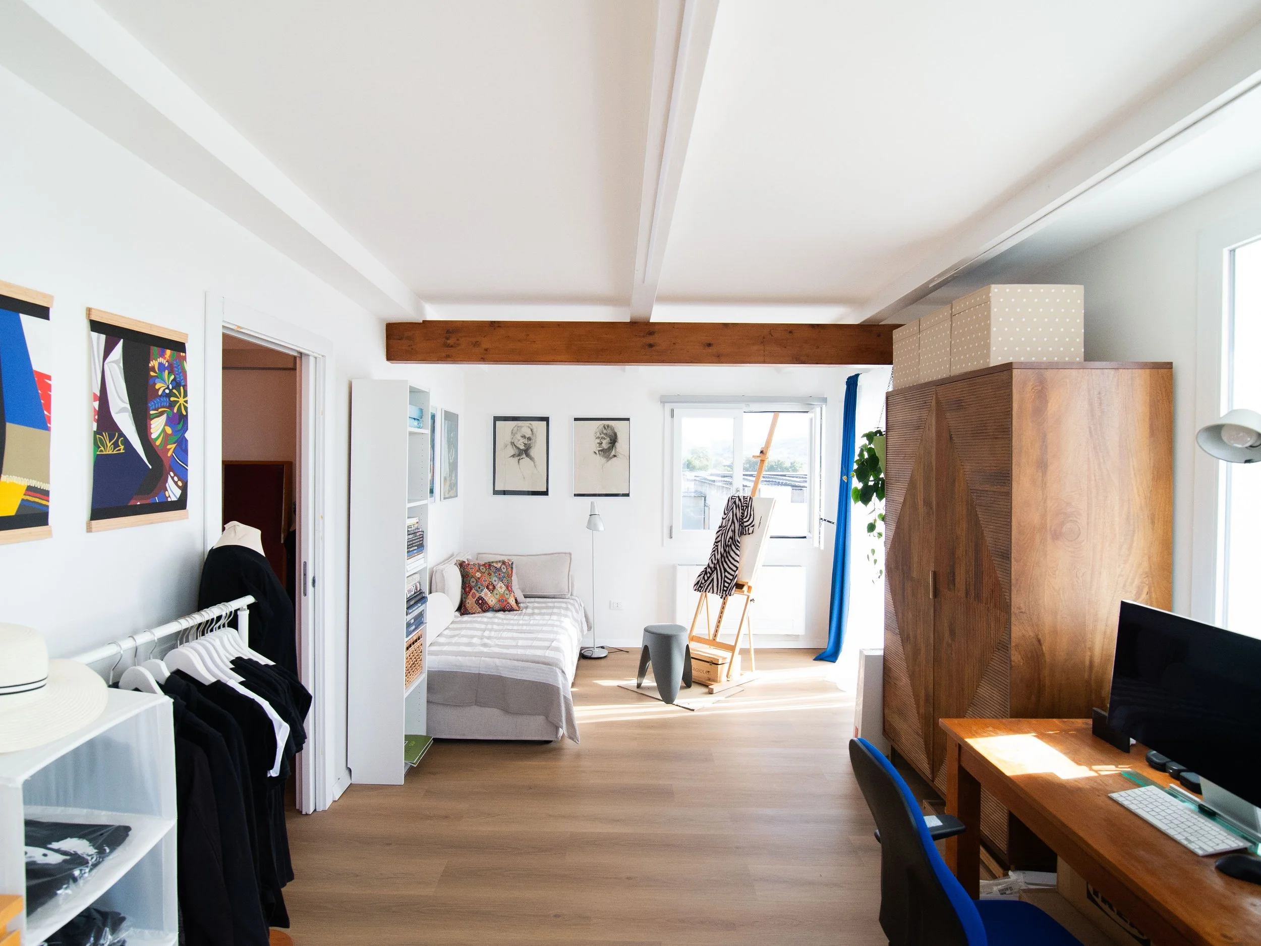 Stanza luminosa con pavimento di legno, letto, libreria con libri, quadro, sedia, armadio in legno, scrivania con computer, pareti biene illuminate da finestre.