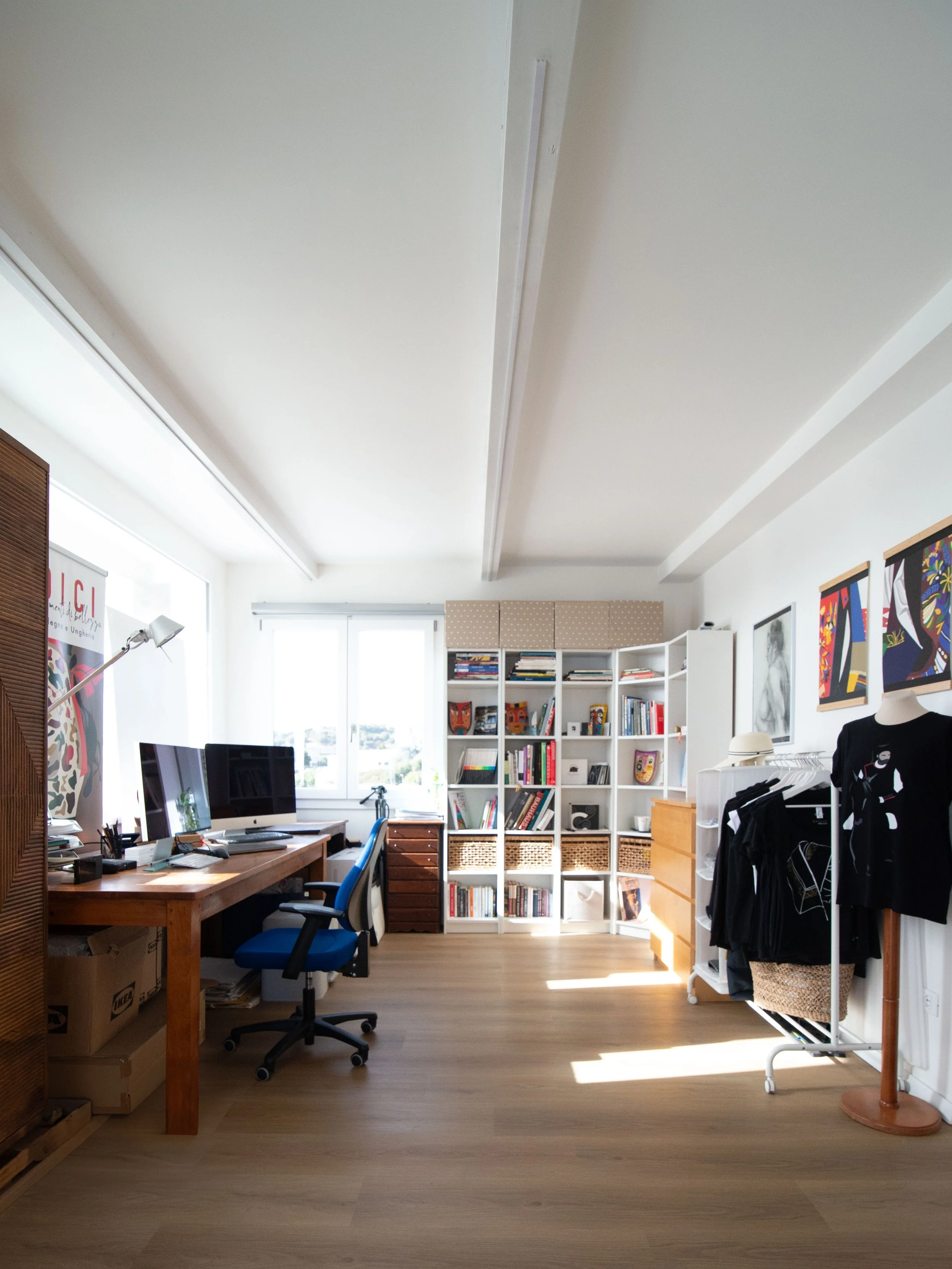 Ufficio arredato con scrivania, due monitor, sedia blu, lampada da scrivania, scaffale con libri e decorazioni, armadio bianco, rack con vestiti, quadri artistici alle pareti, finestre con luce naturale.