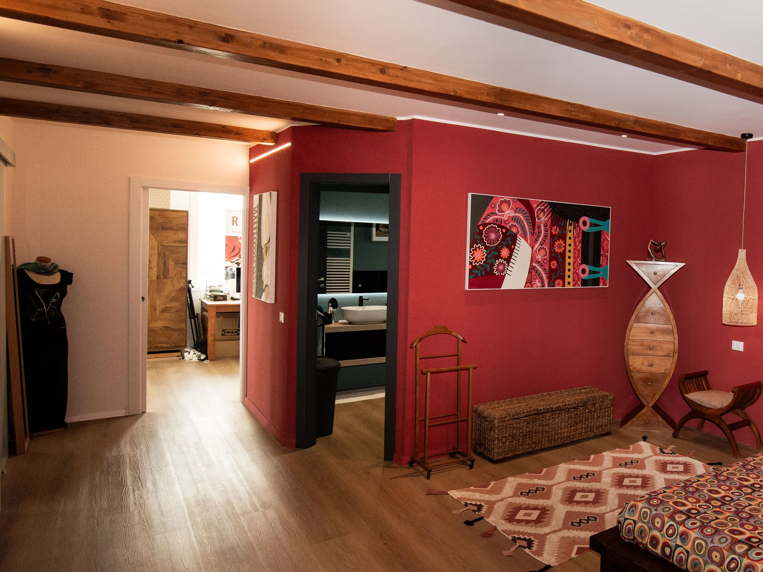 Interno di una stanza con pareti rosse e soffitto con travi di legno. Decorazioni e mobili in stile etnico, con un tappeto con motivi geometrici e un letto con copriletto a colori vivaci.