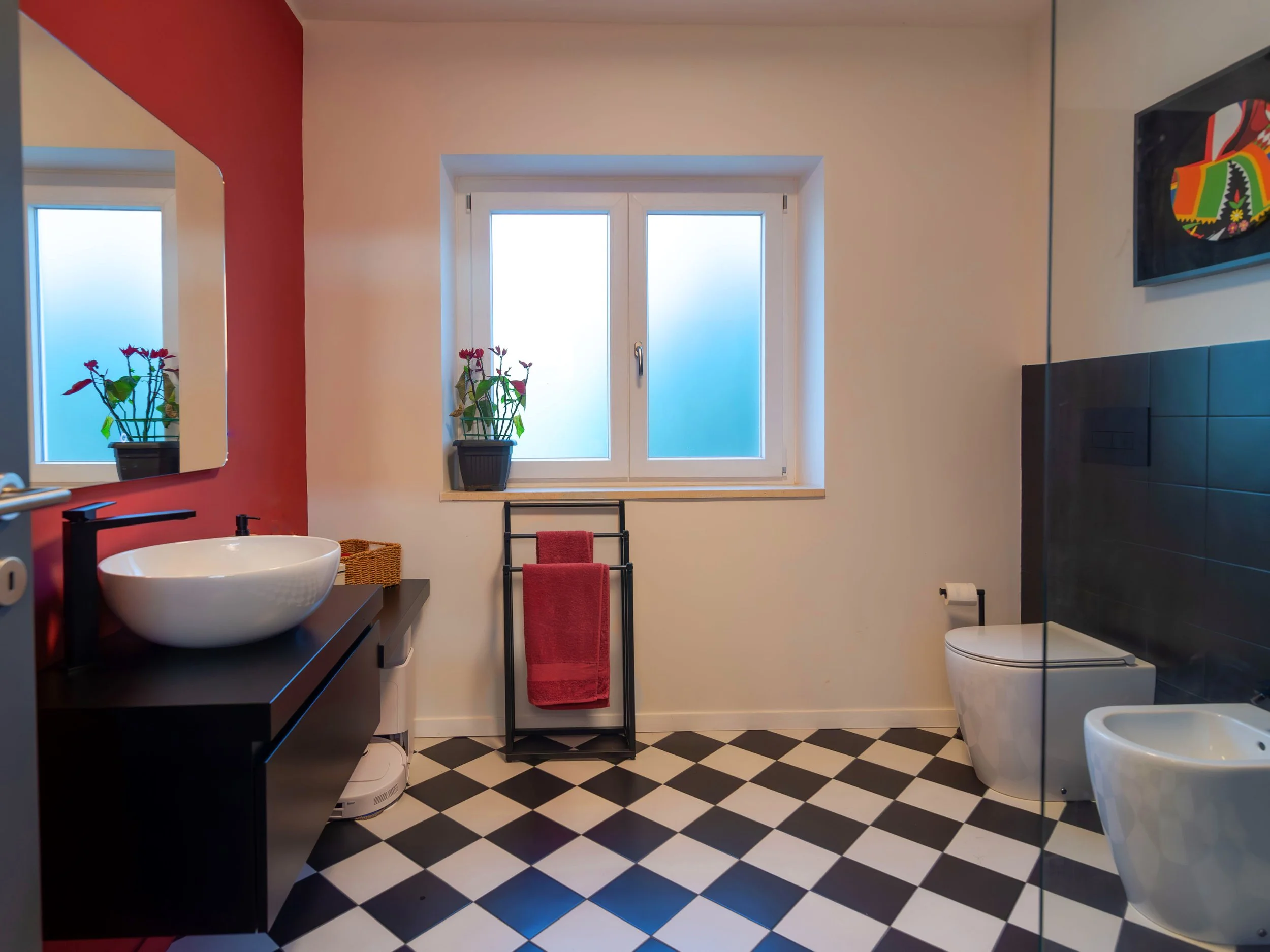 Bagno moderno con lavabo rotondo su mobile nero, specchio, vaso e bidet bianchi, tappeto rossa, piante sul davanzale della finestra, parete rossa, parete bianca, piastrelle a scacchi bianco e nero.