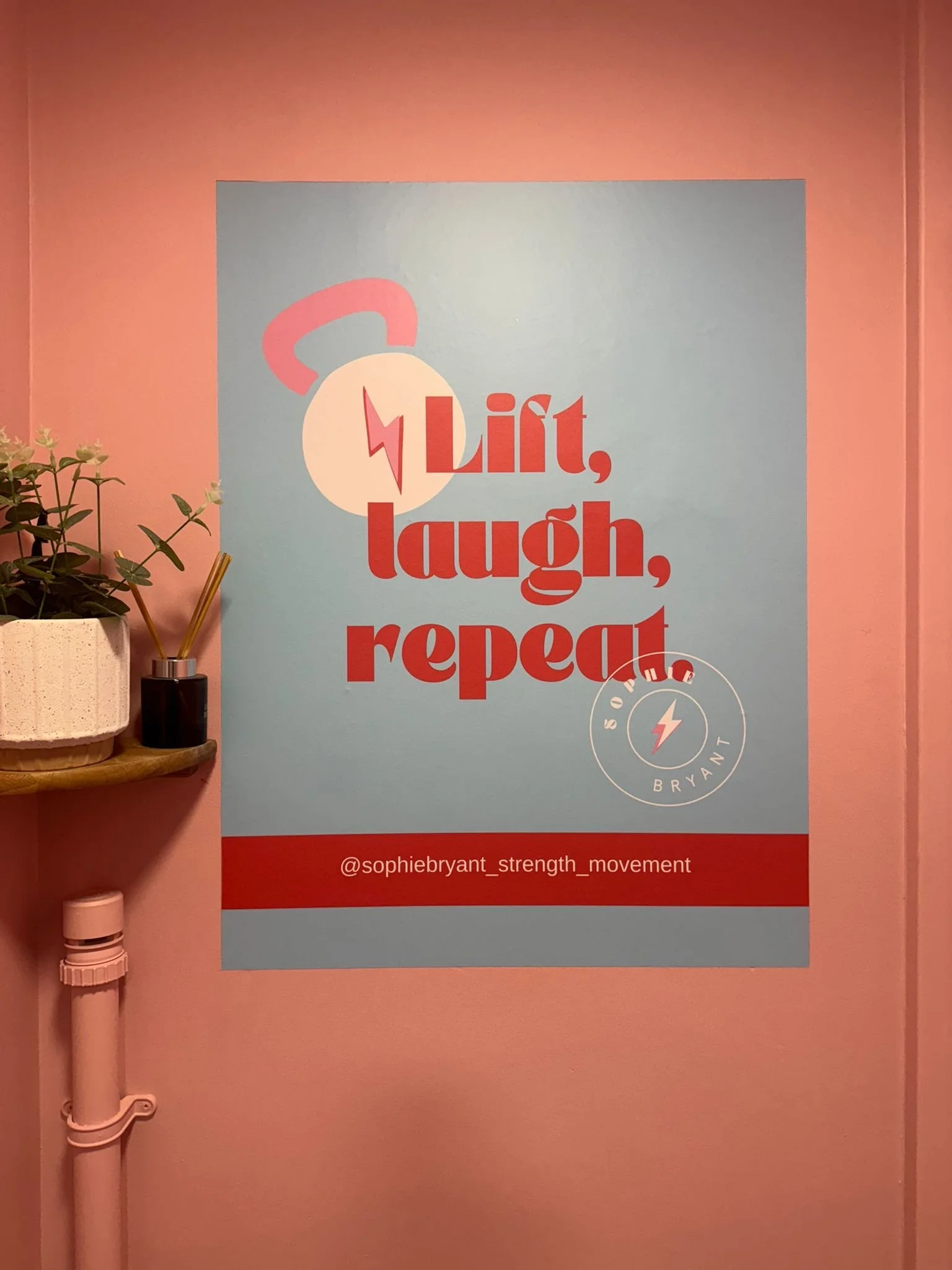 A motivational poster on a pink wall reading 'Lift, laugh, repeat' with a graphic of a kettlebell and a lightning bolt, and an Instagram handle '@sophiebryant_strength_movement'.