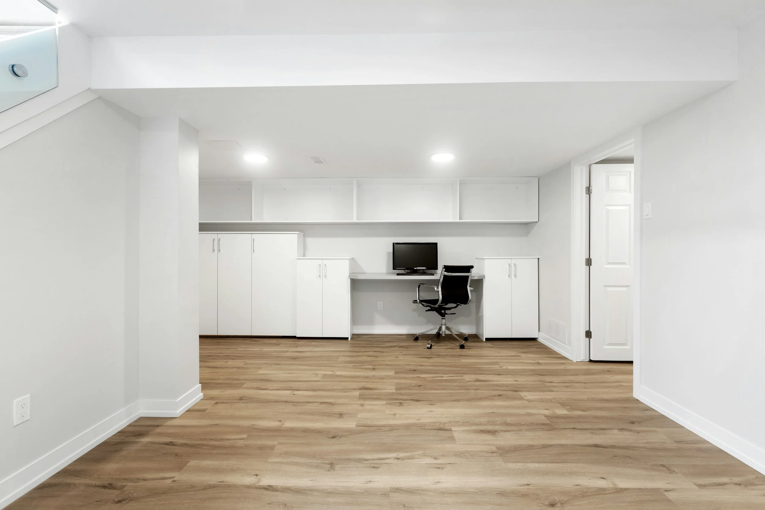 A minimalist home office with white walls, wooden floor, built-in white cabinets, a small desk with a computer monitor, and a black office chair.